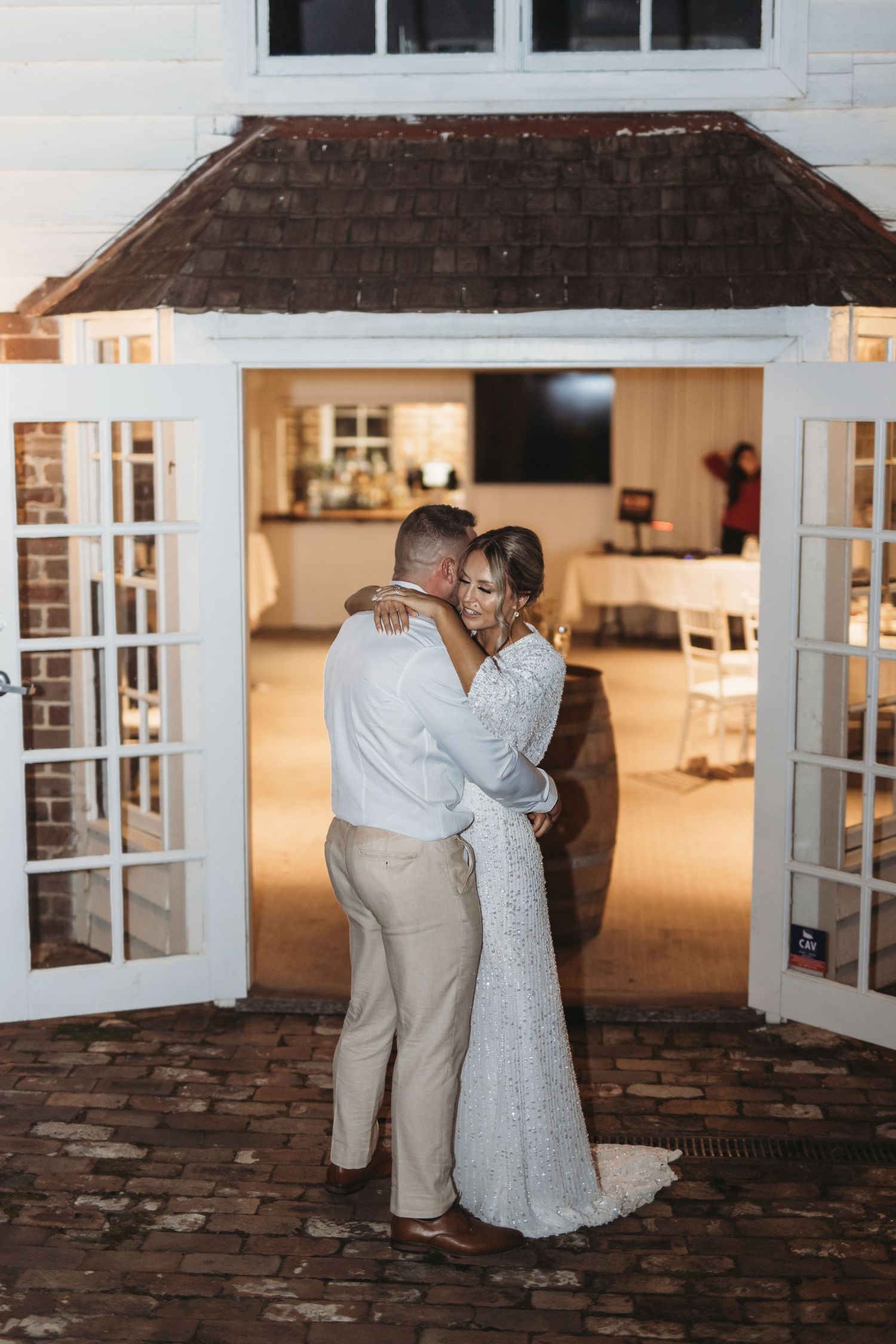 Romantic wedding couple dancing in doorway of illuminated white building at dusk.