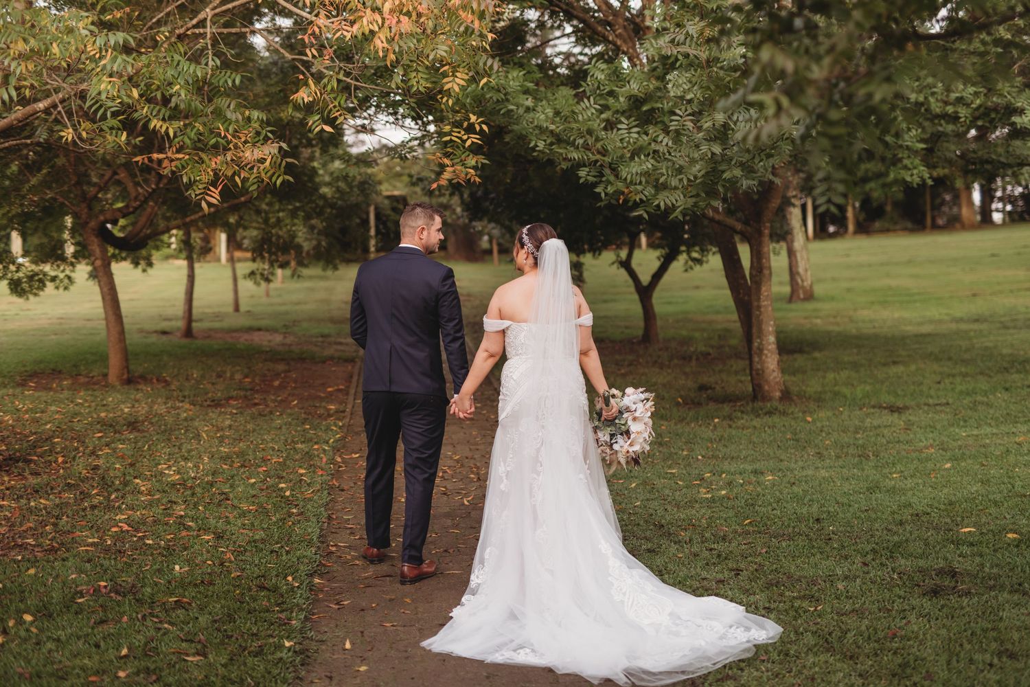 A wedding couple walks hand in hand down a tree-lined path in an autumn setting.