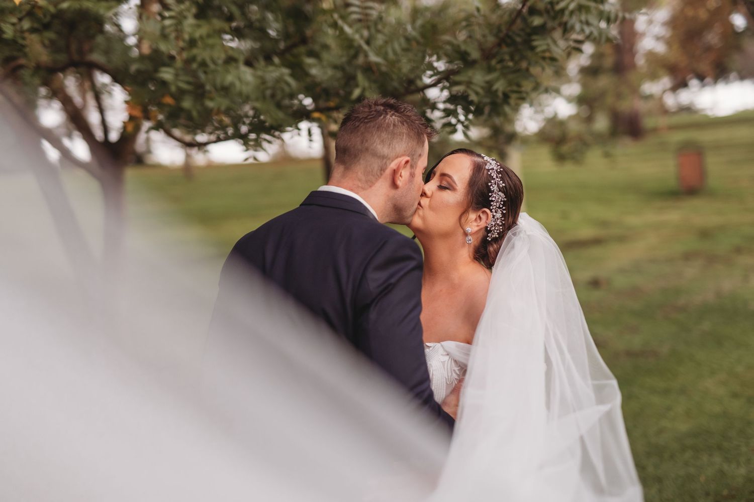 A romantic wedding kiss captured outdoors in a park setting with soft natural lighting.