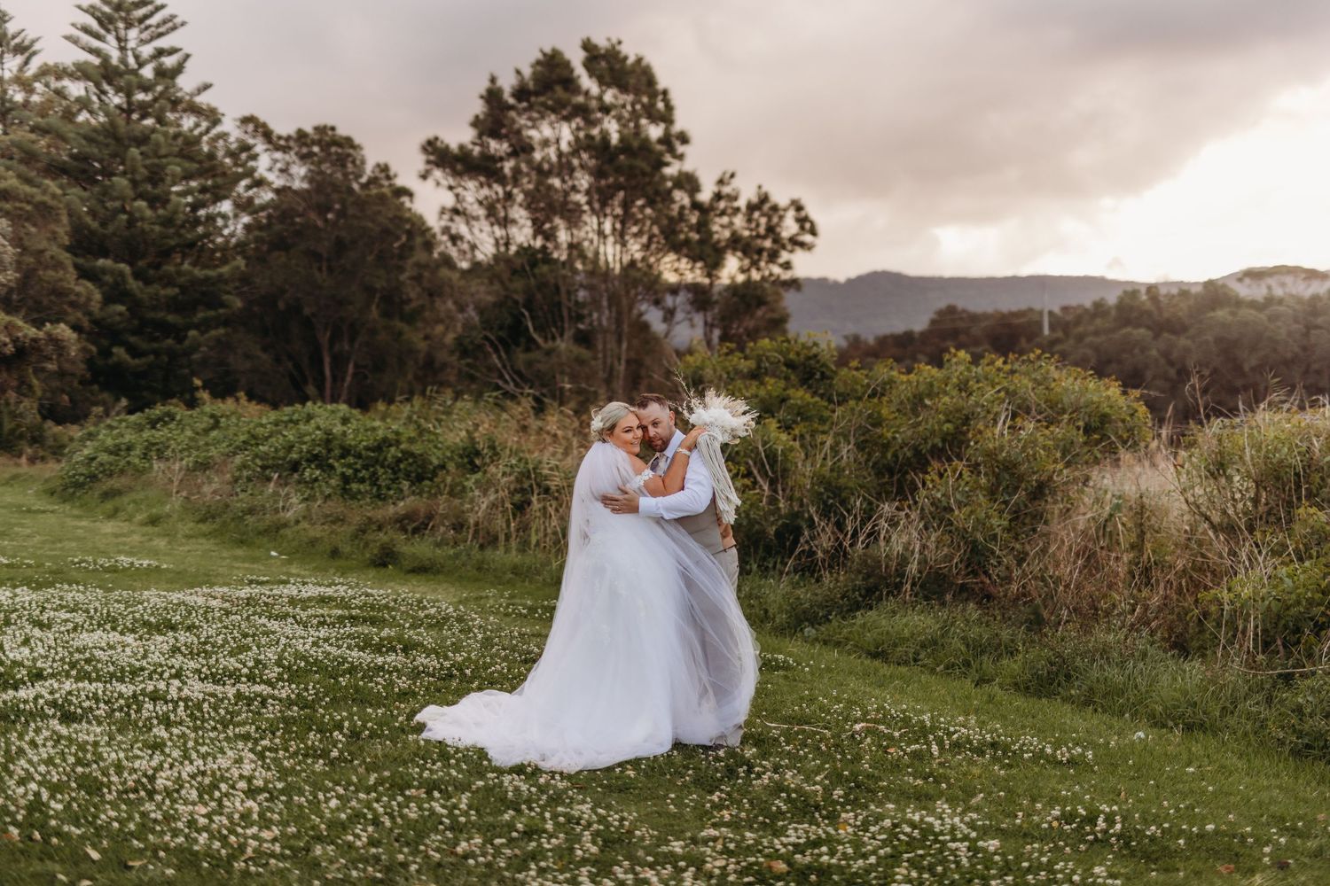 A romantic wedding couple embraces on a hilltop overlooking mountains at dusk.