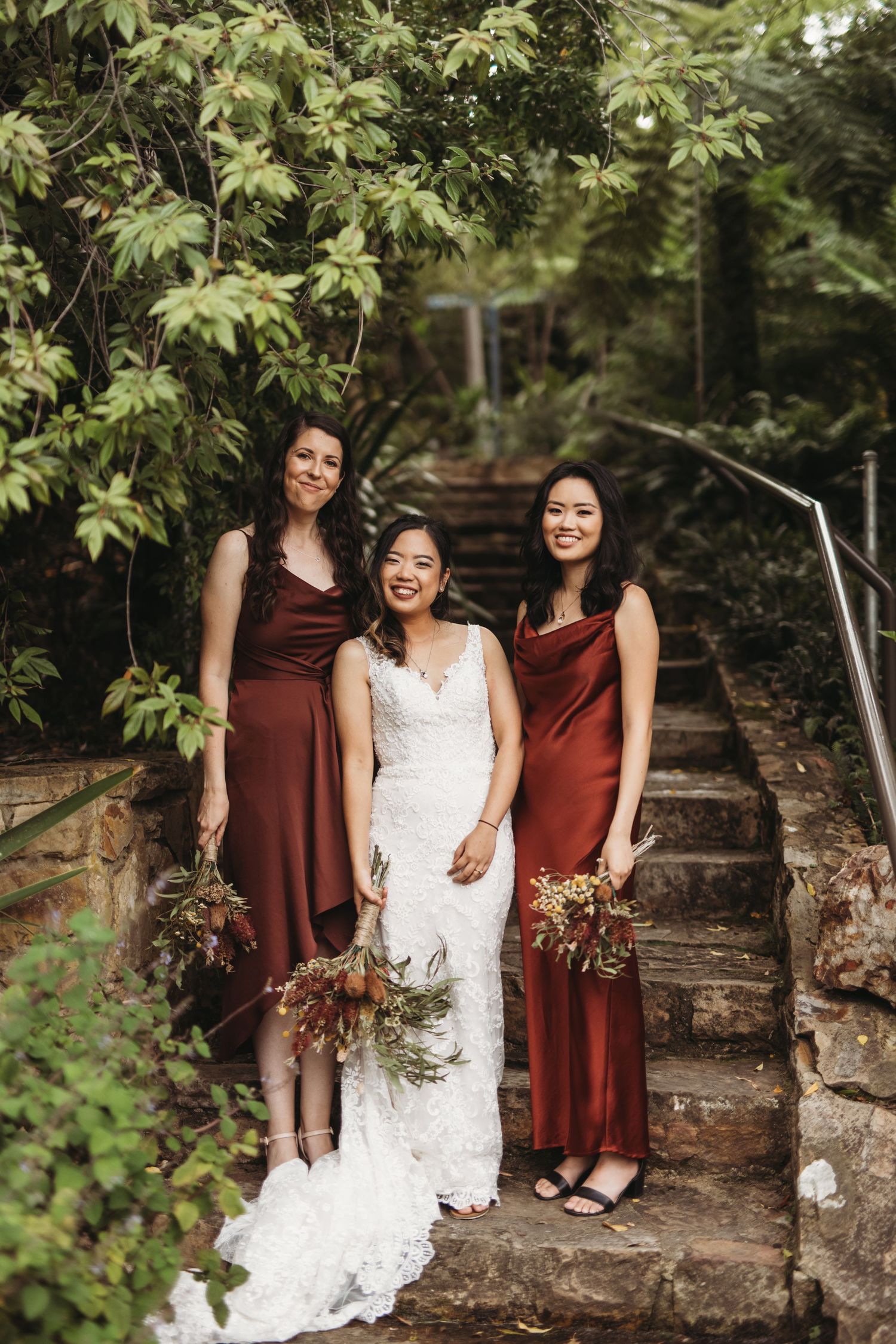 Bridesmaids in burgundy dresses pose with bride on rustic stone steps surrounded by foliage.