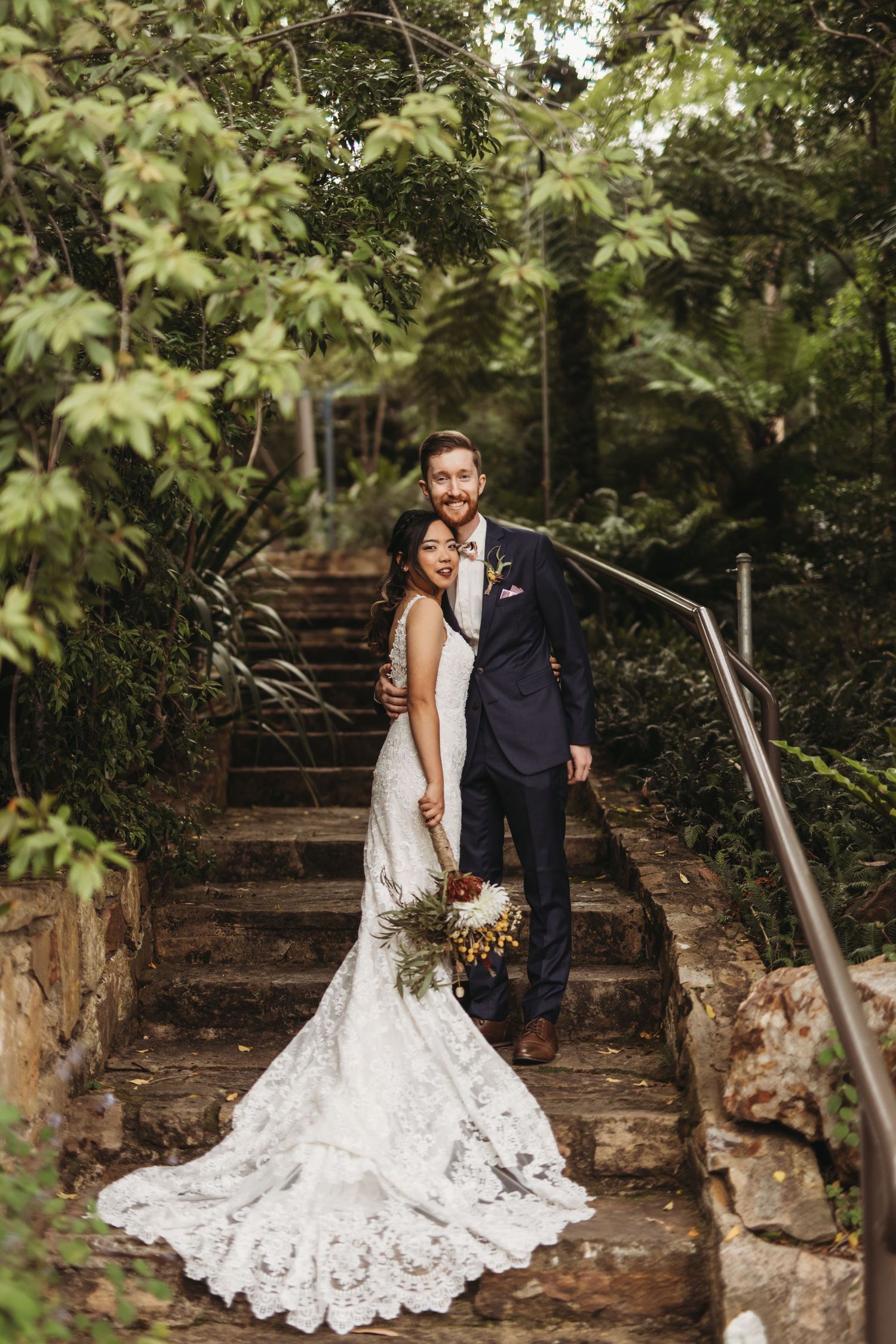 A couple poses on stone garden steps with flowing white wedding dress train amid lush greenery.