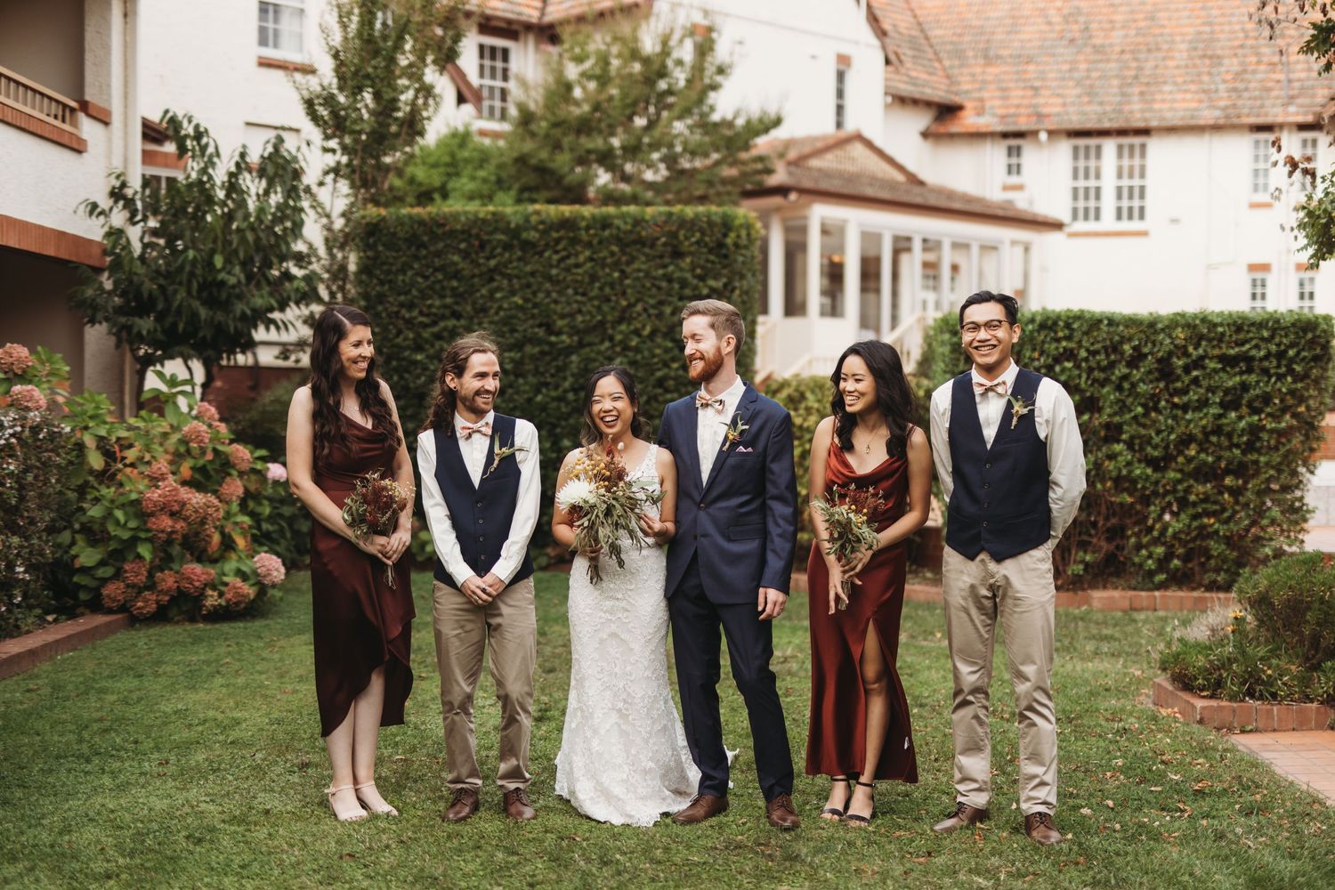 Wedding party poses together on a lawn in front of a stately house with manicured hedges.