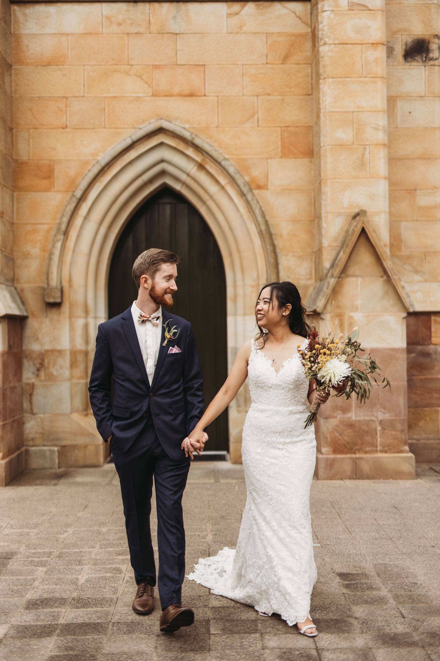 A happy couple walks hand in hand in front of a historic stone church entrance.