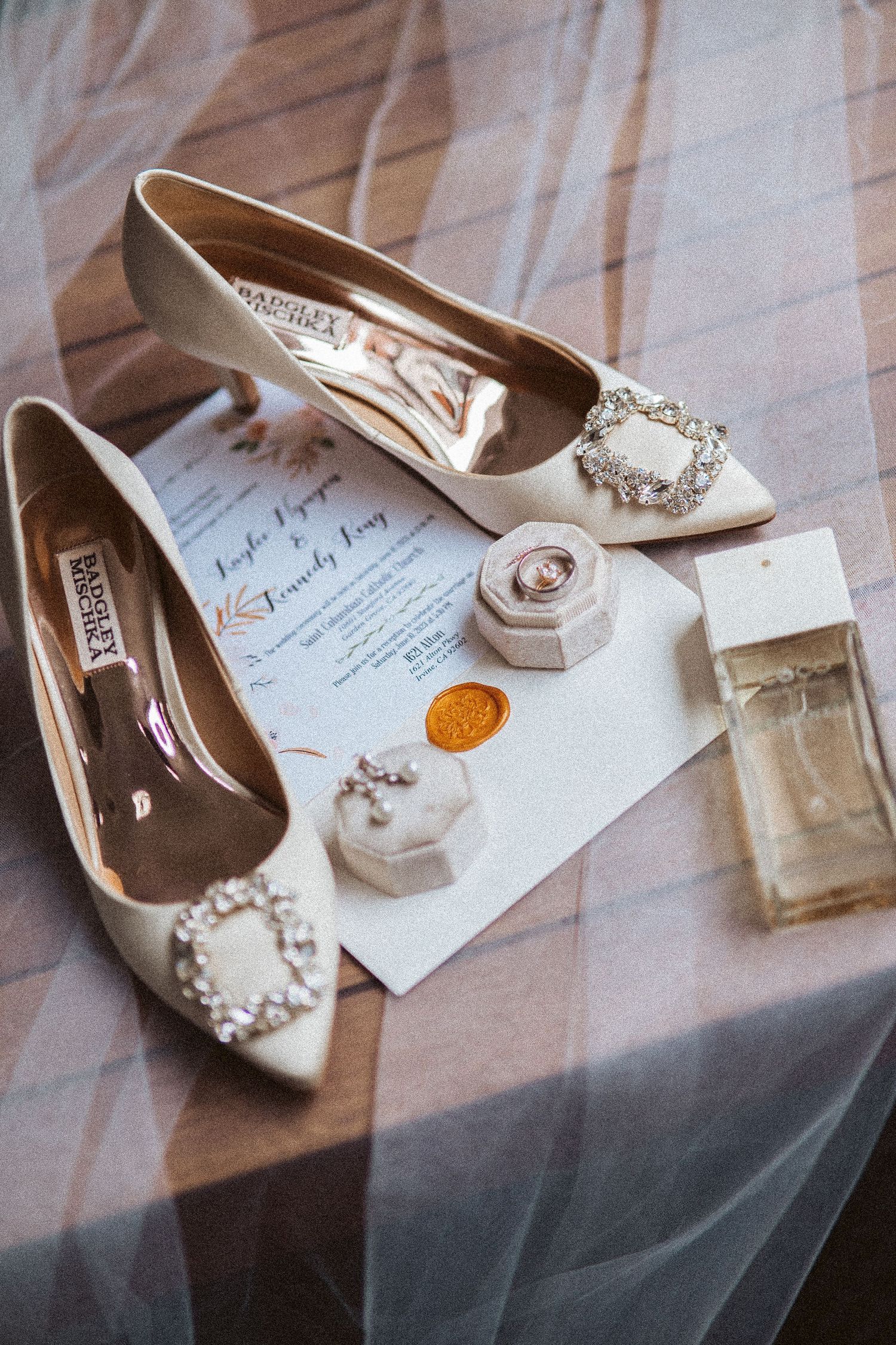 Wedding shoes, perfume bottle and rings arranged with invitation on light fabric.
