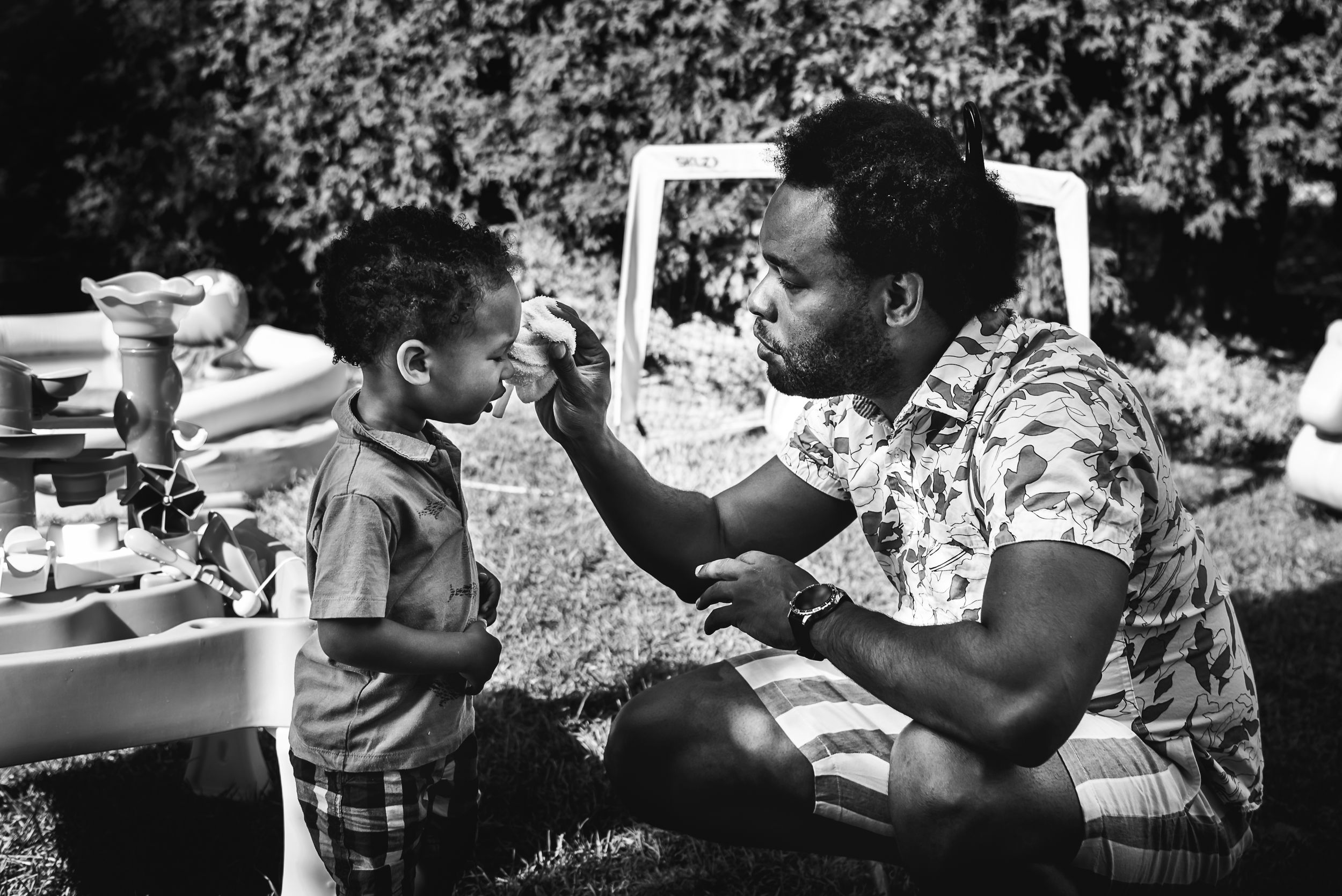 BW image of a black young child having his tears cleaned off by his father, who crouches in front of him.