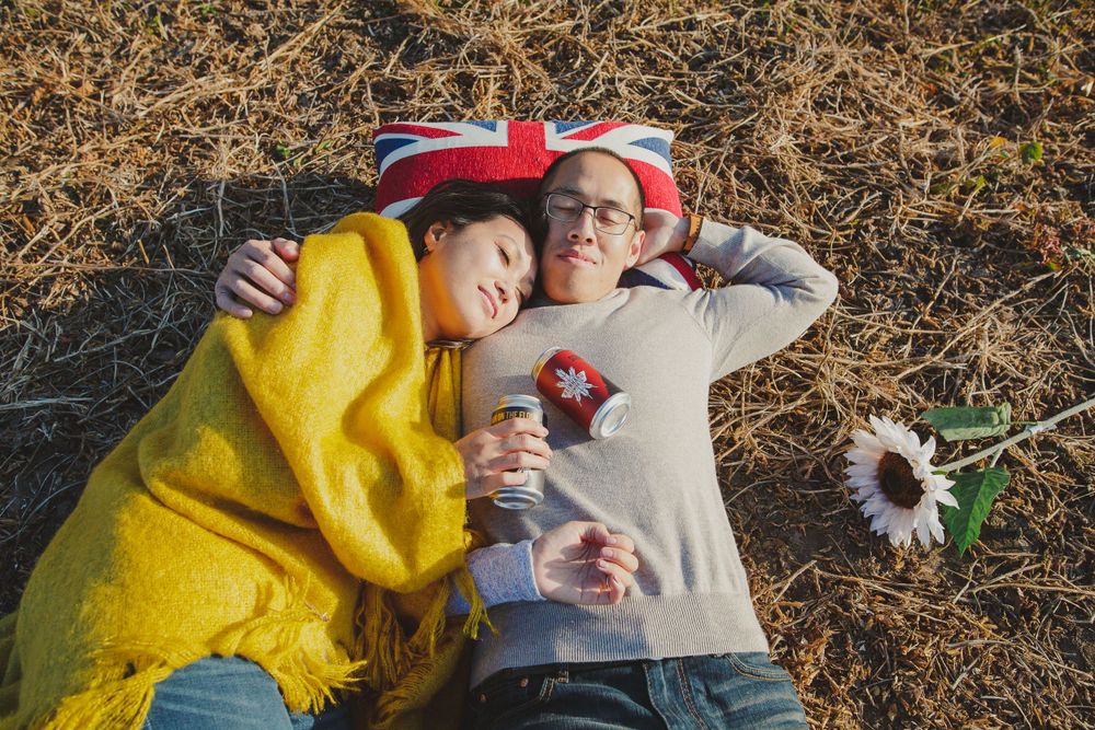 A couple relaxes on dry grass wearing casual attire with a flower nearby.