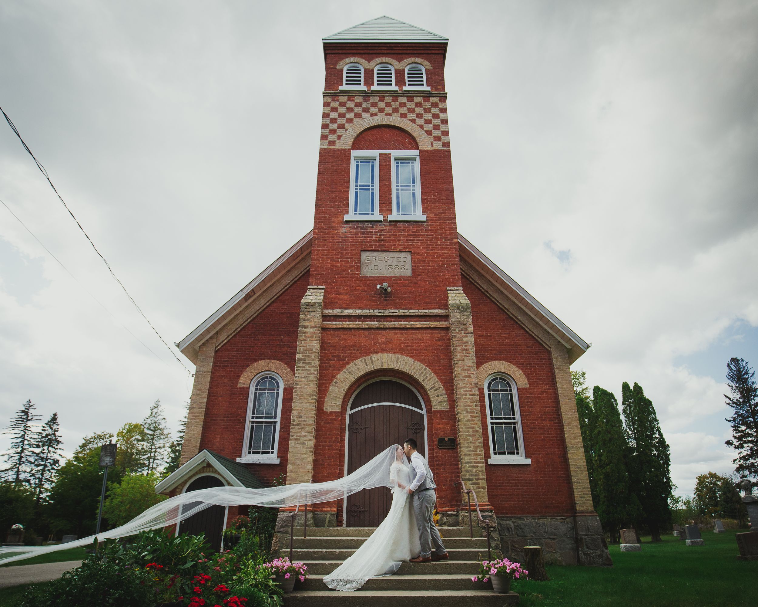 A historic red brick church with tall tower serves as backdrop for a bride on its steps.