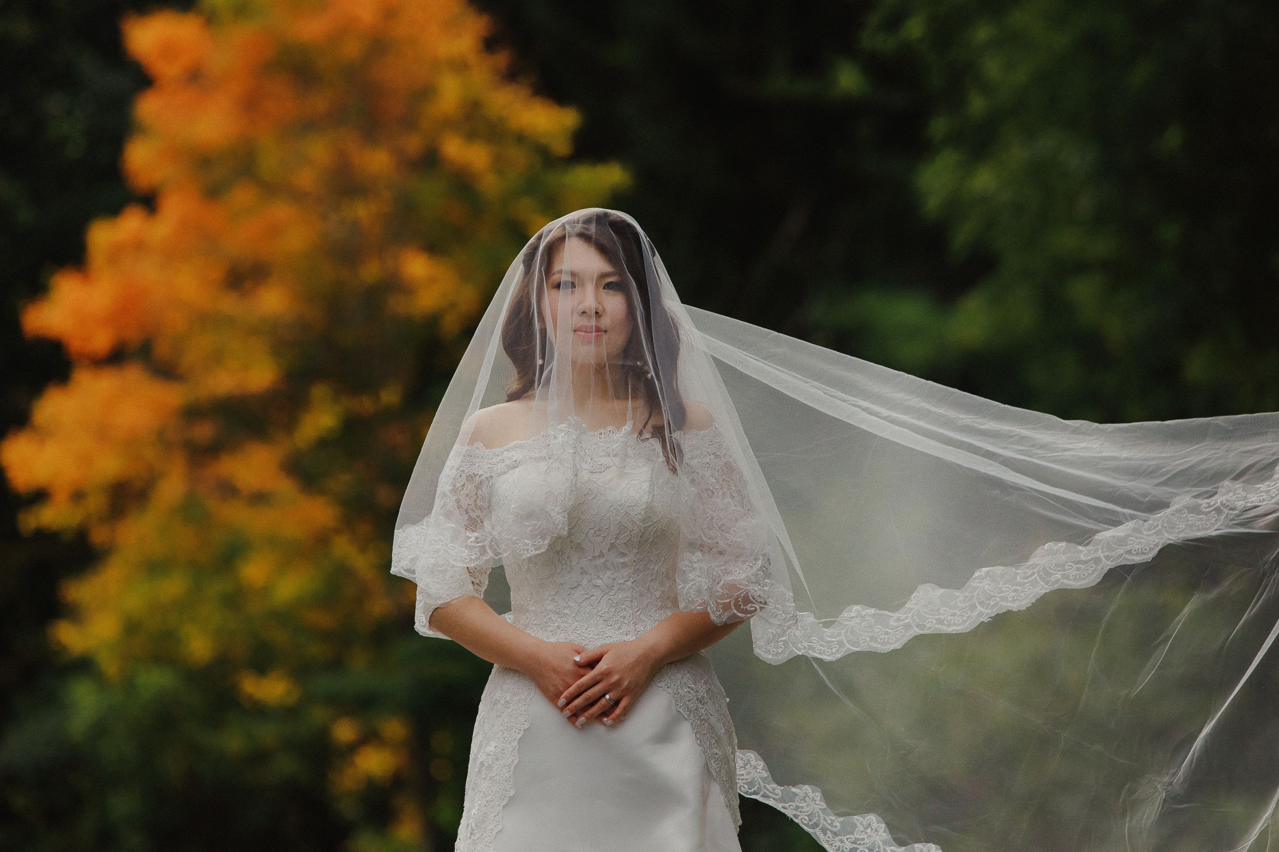 A bride in a lace wedding dress with flowing veil poses against vibrant fall foliage.