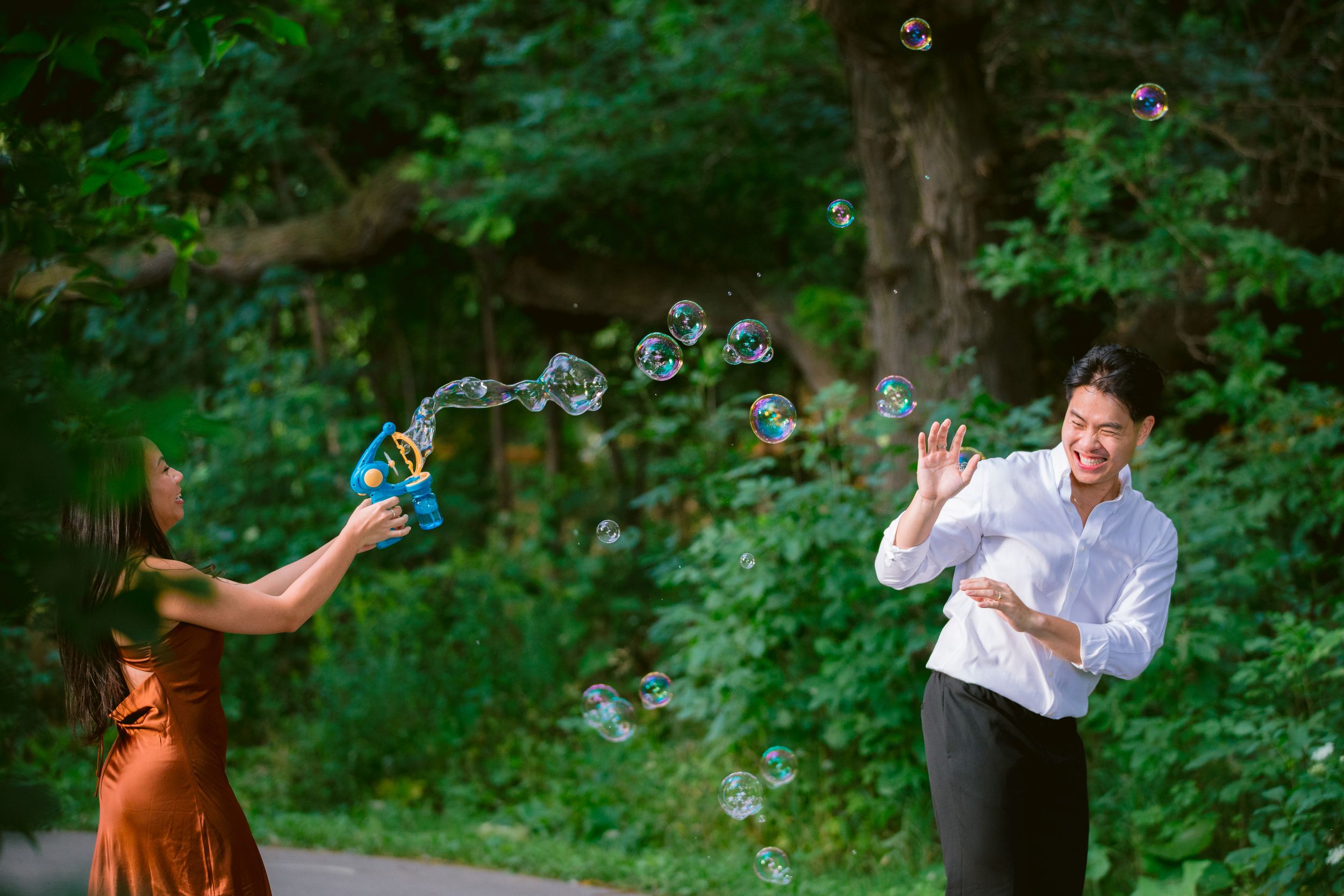 A playful couple blows bubbles at each other on a tree-lined path in a lush green setting.
