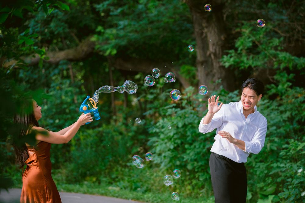 A playful couple blows bubbles at each other on a tree-lined path in a lush green setting.