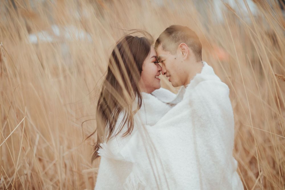 A couple shares an intimate moment wrapped in white blankets among tall wheat grass.