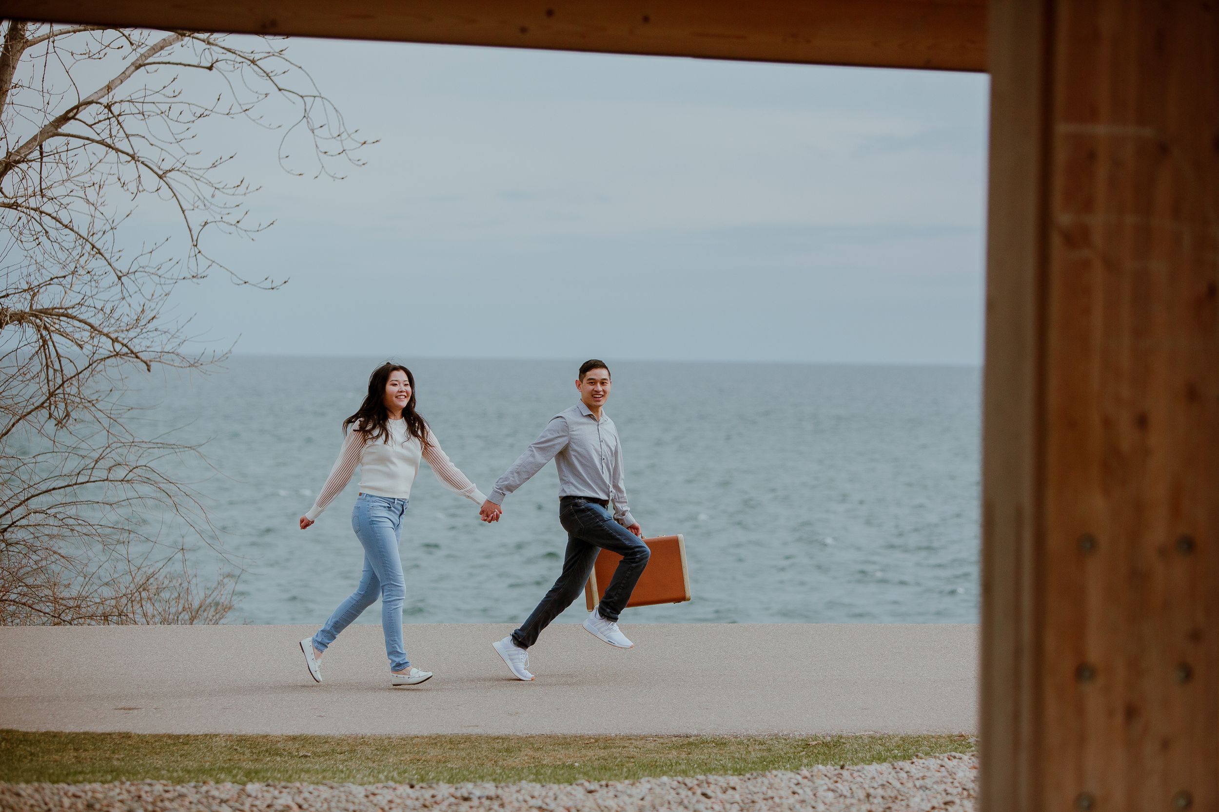 A couple runs joyfully along the waterfront while holding hands with one carrying a brown suitcase.