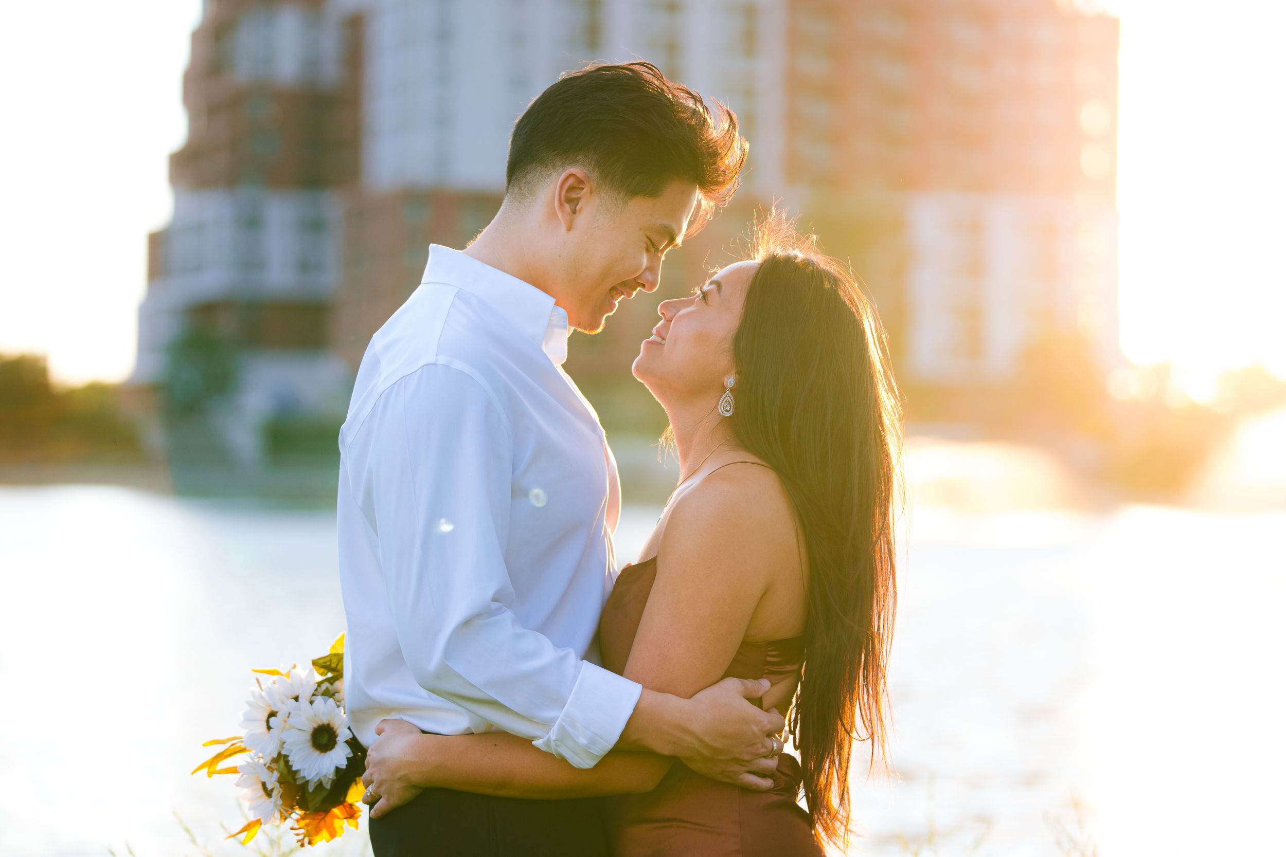 A tender moment between a couple backlit by warm sunset glow near a waterfront.