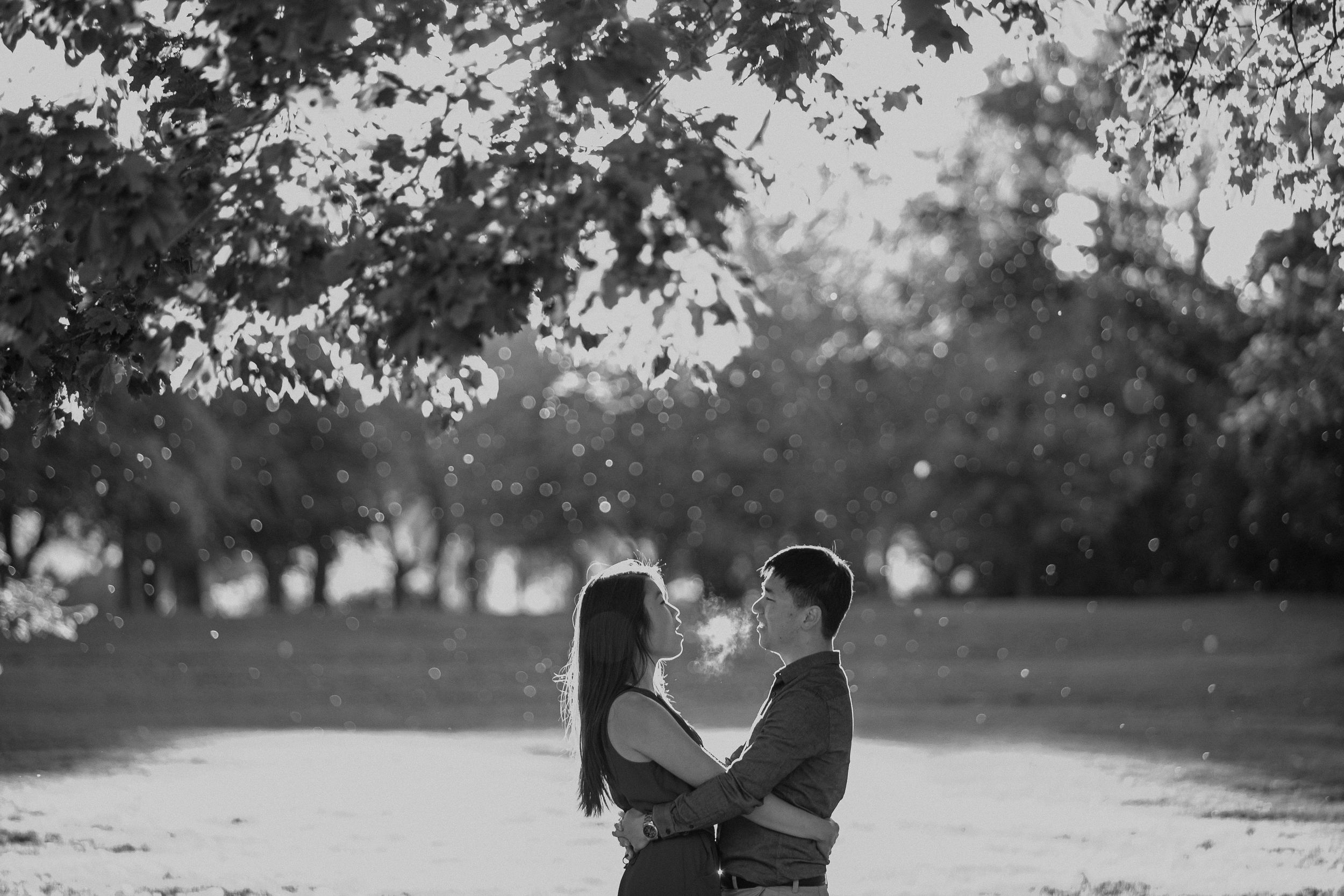 A romantic black and white portrait of a couple embracing under tree branches.