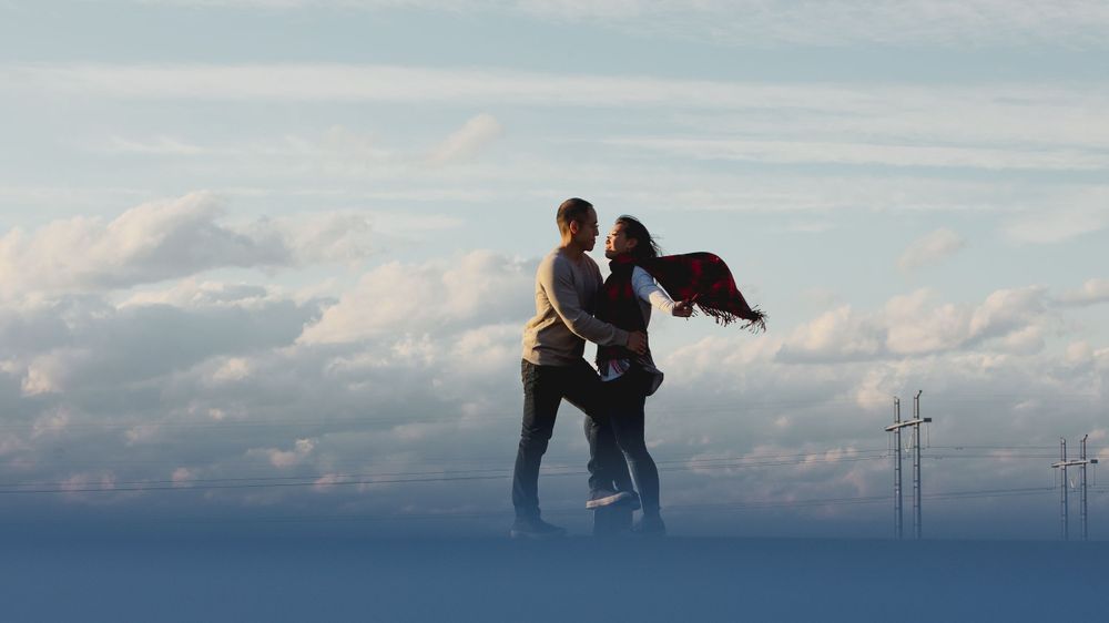 A couple poses against cloudy skies with wind turbines in the distance.
