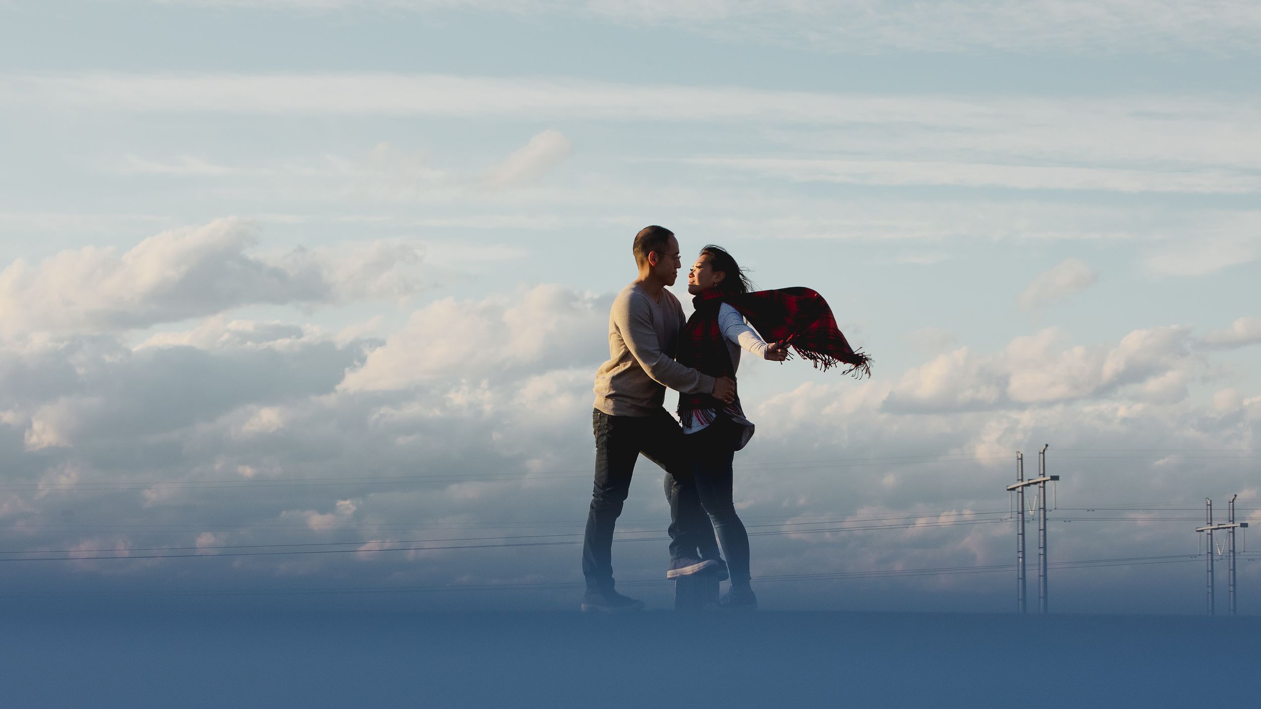 A couple poses against cloudy skies with wind turbines in the distance.