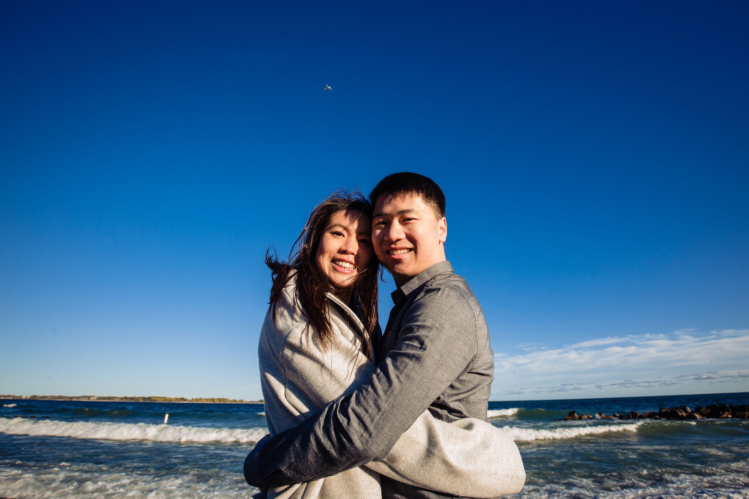 A loving embrace shared at the ocean with blue skies and waves in the background.