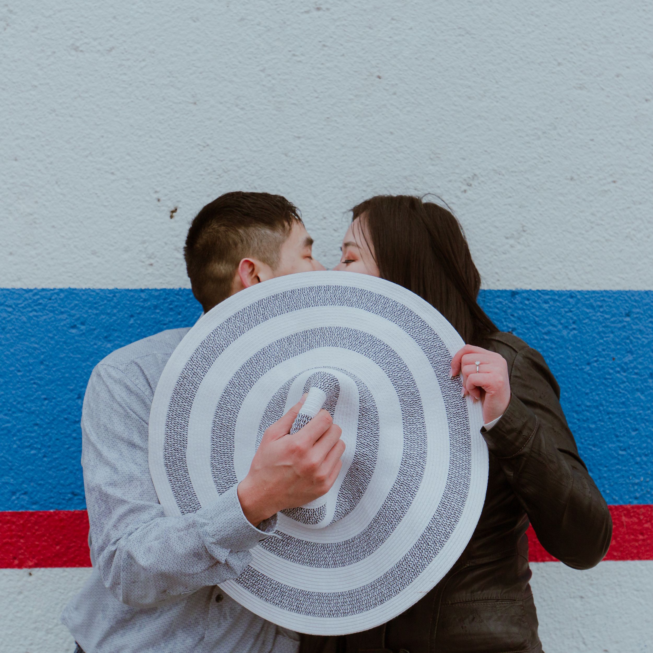 A couple shares a kiss behind a circular striped disc against a blue and white wall.