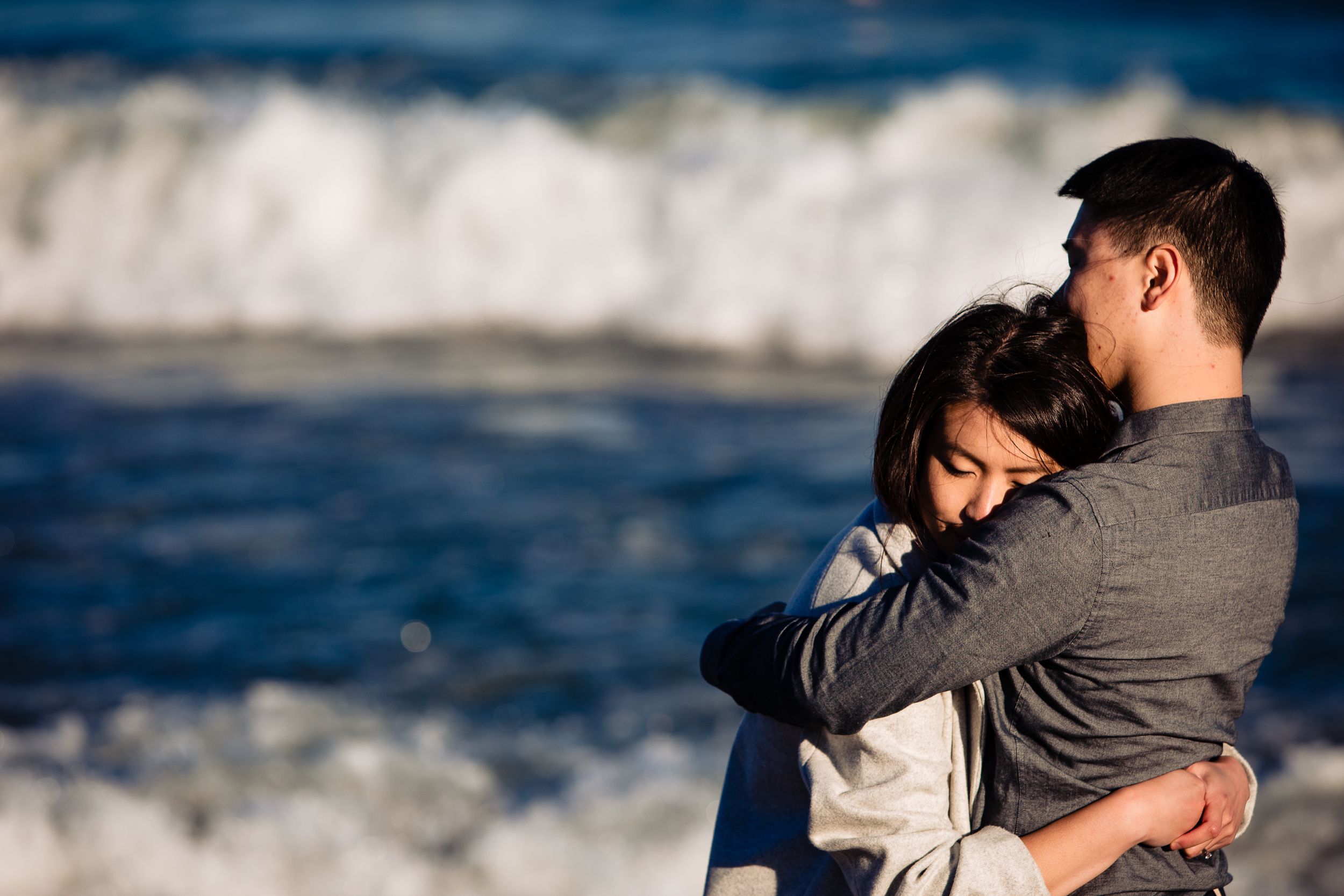 A tender embrace by the ocean with white waves crashing in the background.