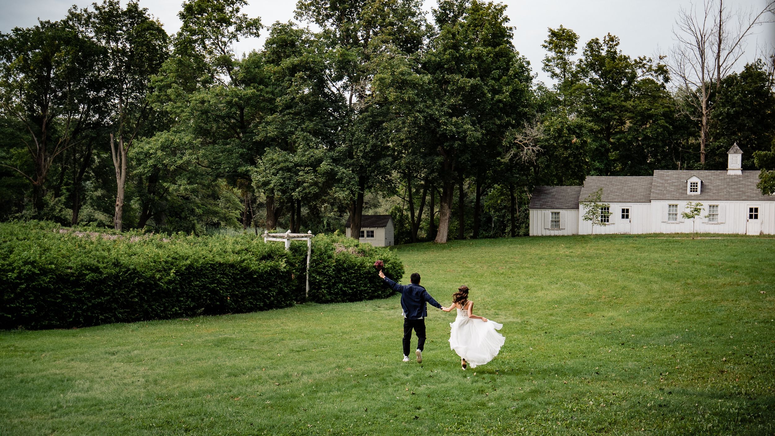 Couple in wedding attire walking across a scenic lawn with a white house in background.