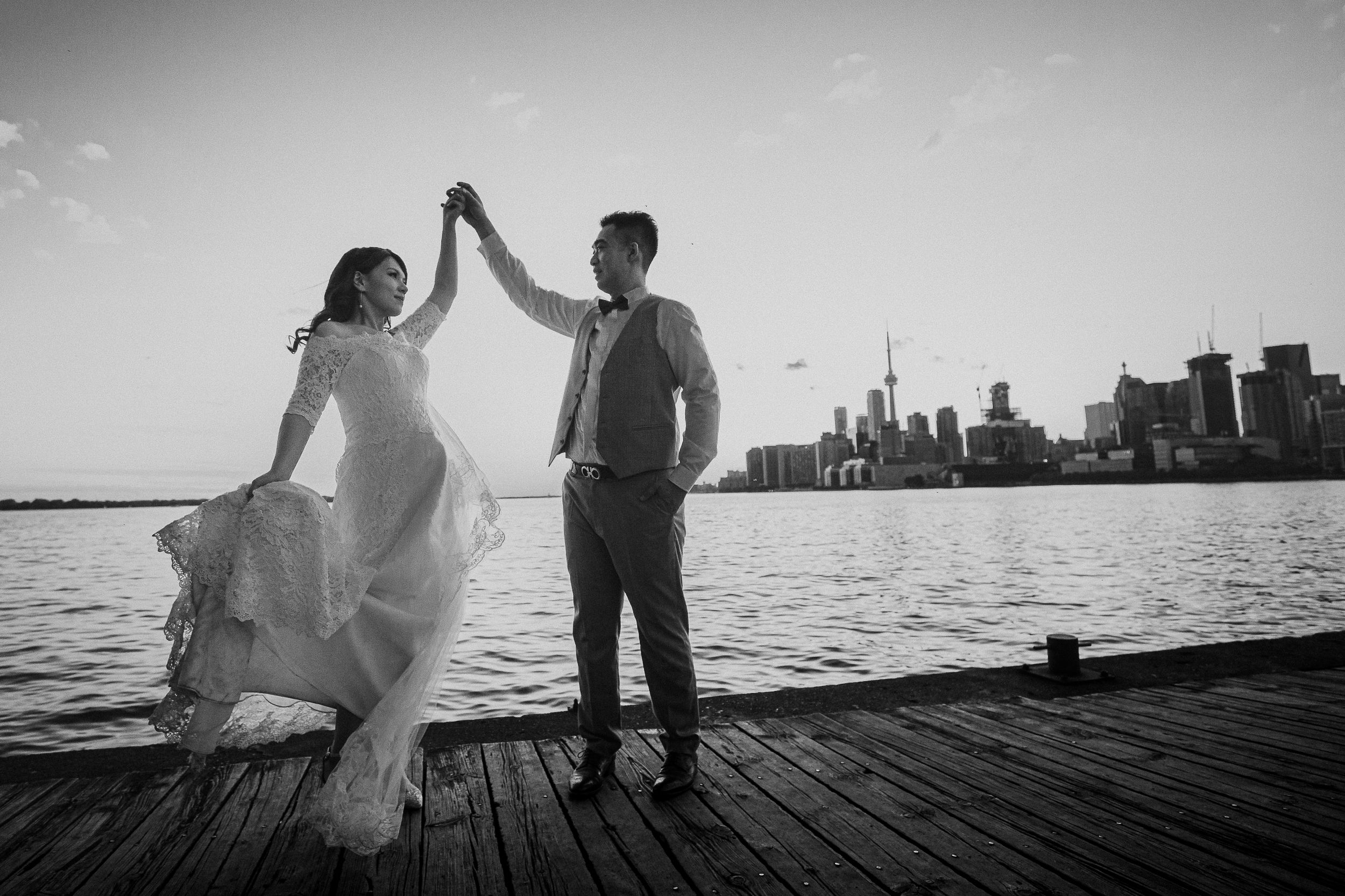 Black and white photo of couple dancing on pier with city skyline in background.