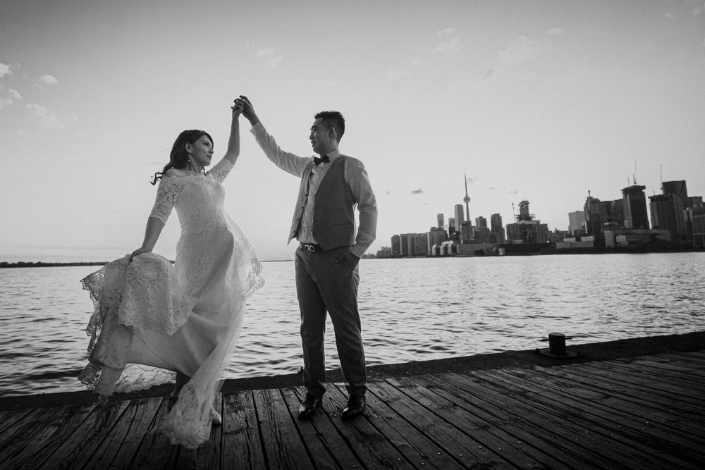 Black and white photo of couple dancing on pier with city skyline in background.
