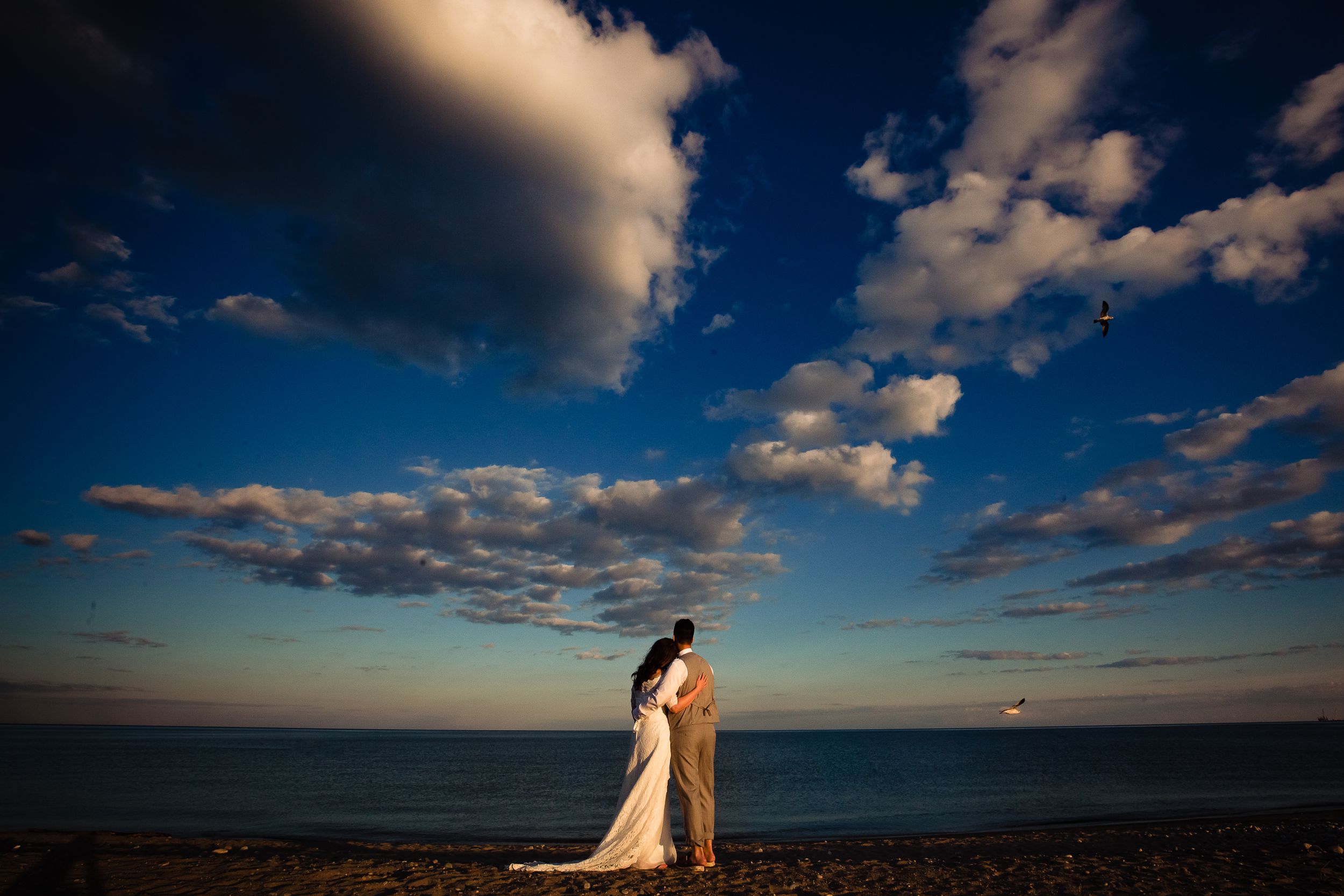 Couple embraces on beach at sunset with dramatic clouds in deep blue sky.