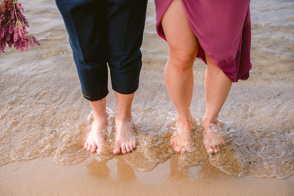 Two pairs of bare feet standing in shallow ocean waves on a sandy beach.