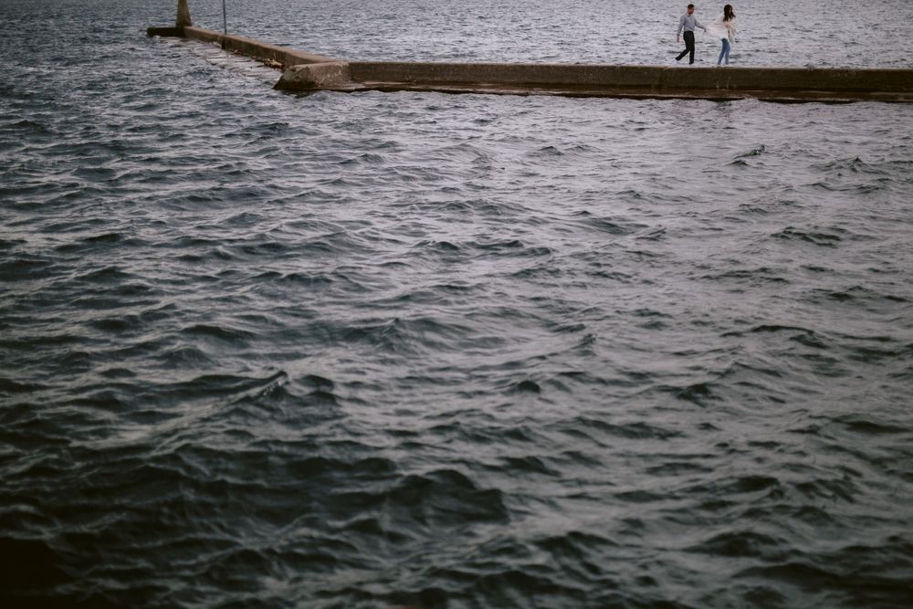 Two silhouettes stand on a pier extending into rippling water.