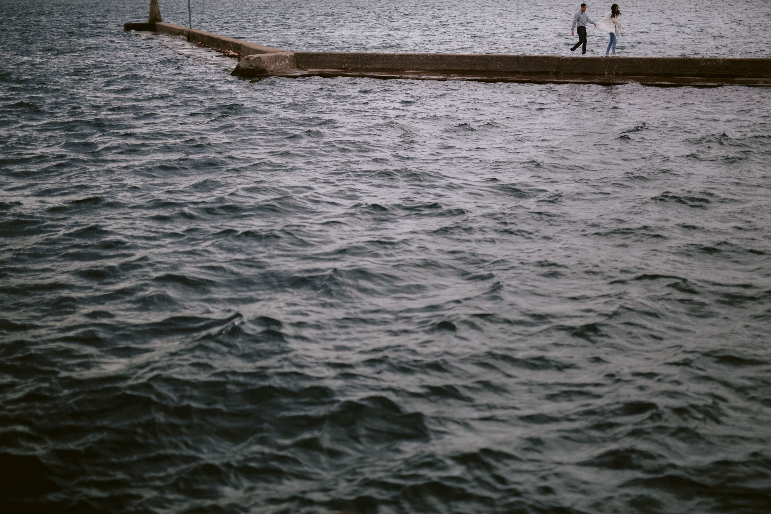Two silhouettes stand on a pier extending into rippling water.
