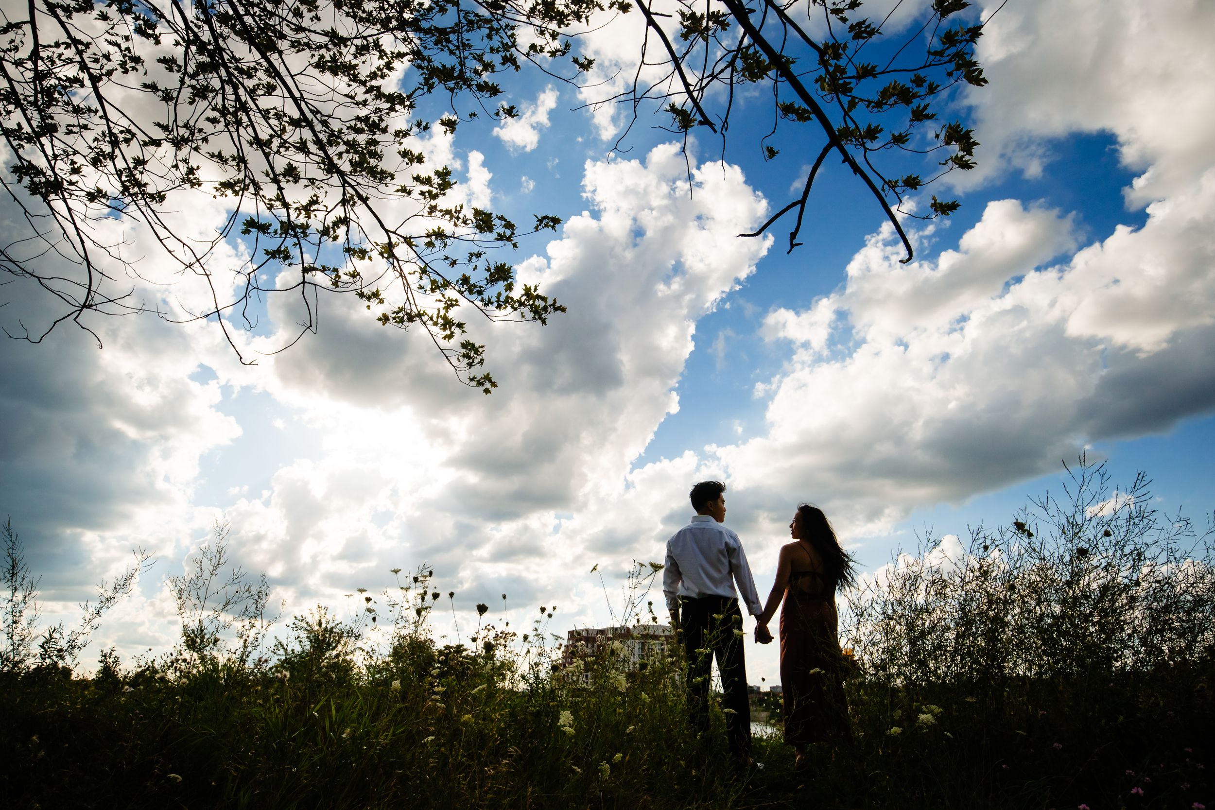 A couple stands hand in hand on a hilltop silhouetted against a dramatic sky with billowing clouds.