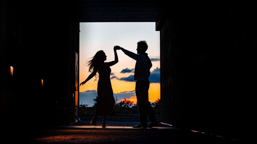 Silhouettes of two people creating a heart shape with their arms against a sunset sky through a doorway.