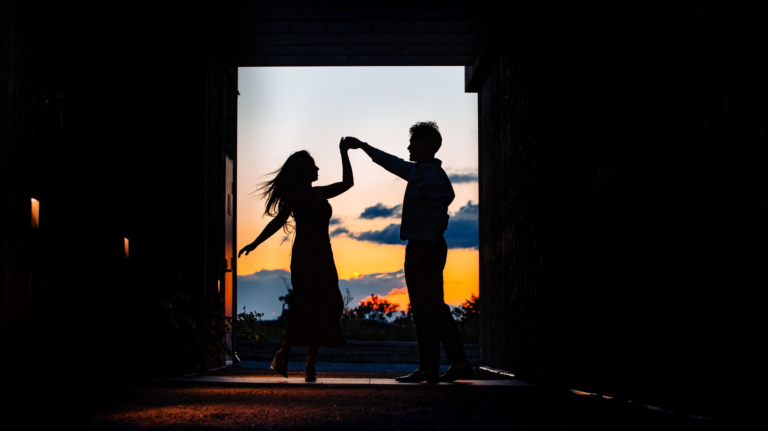 Silhouettes of two people creating a heart shape with their arms against a sunset sky through a doorway.