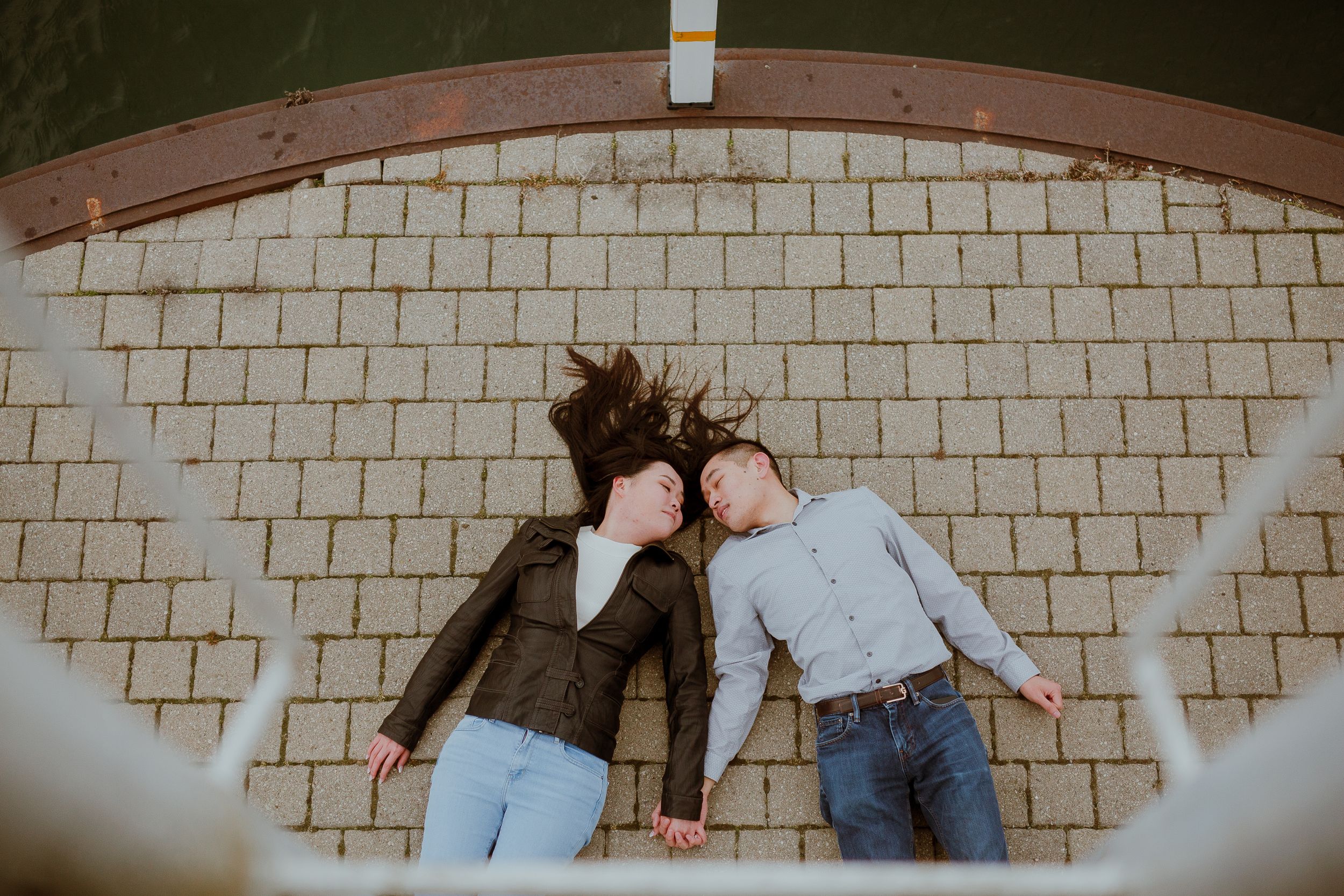 A playful couple lies head-to-head on brick pavement, shot from above.