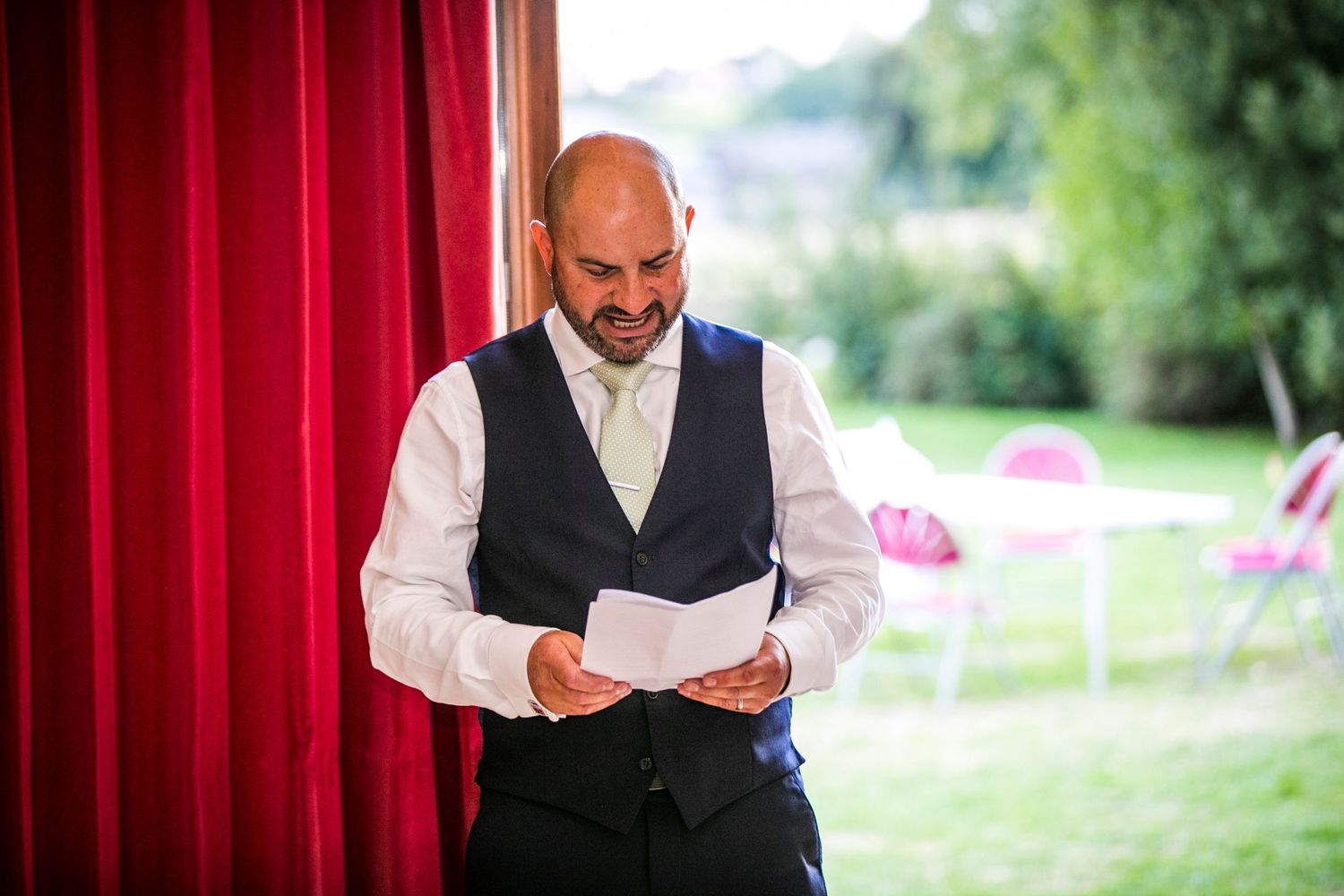A speaker in a vest and white shirt reads from papers near red curtains at an outdoor venue.
