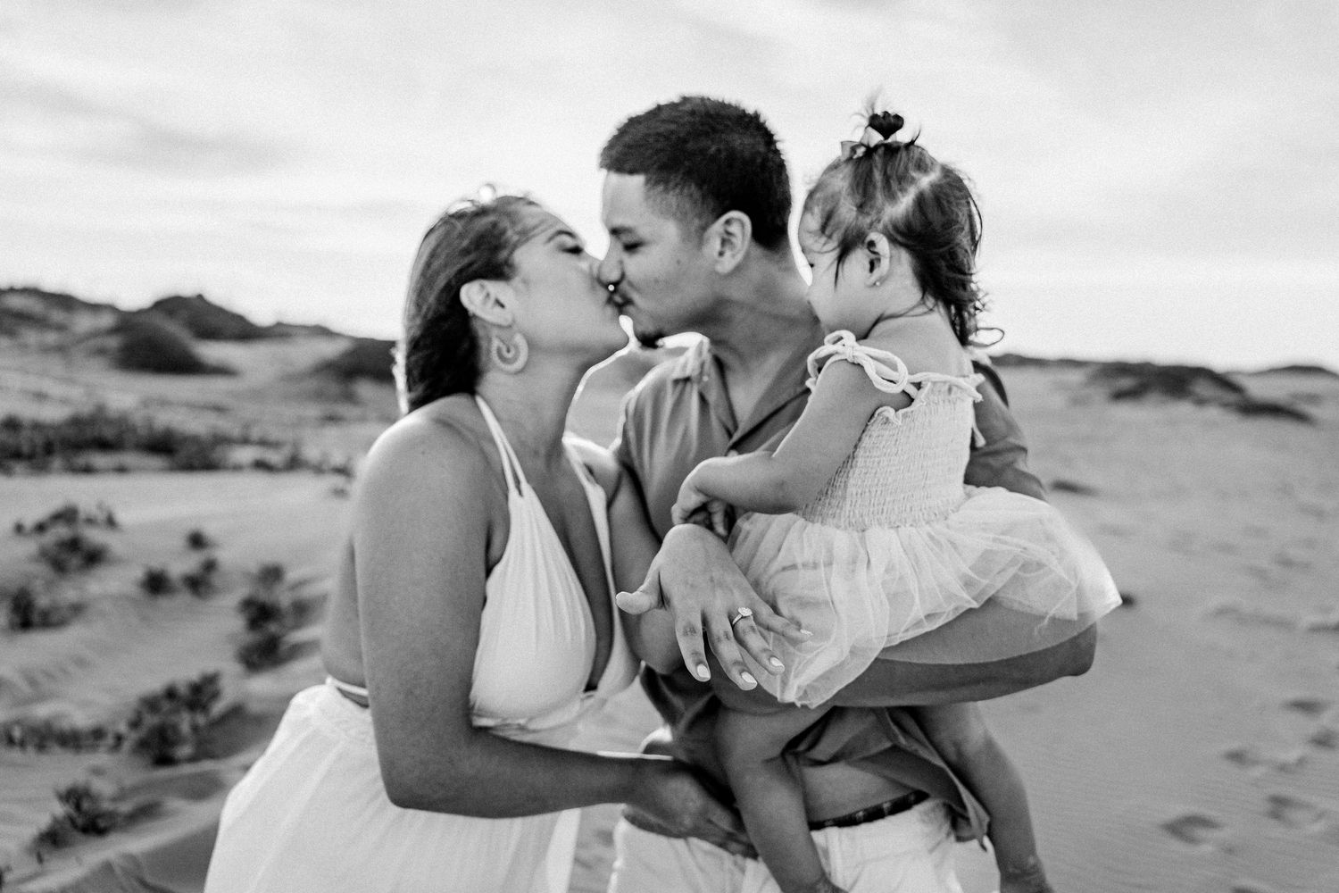 Black and white photo of a couple kissing while holding their toddler daughter on sandy desert dunes.