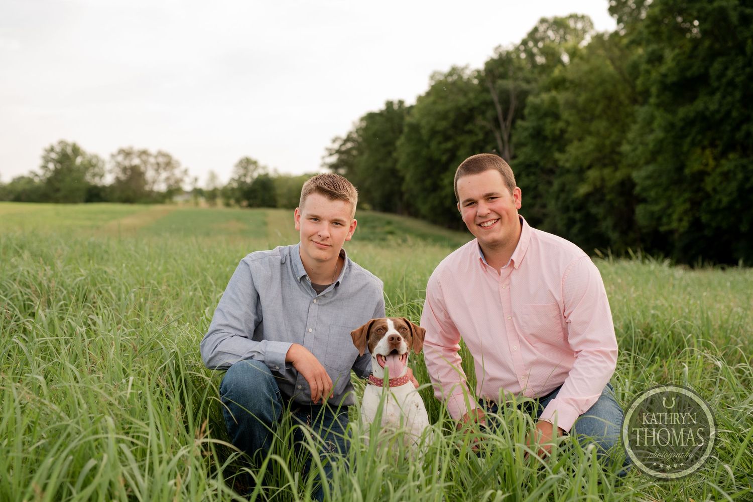 Two people sitting with small dog in green field.