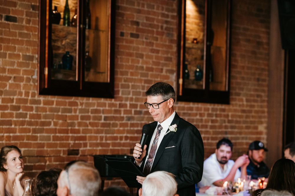 Man delivers wedding speech into microphone at reception in rustic brick-walled venue.