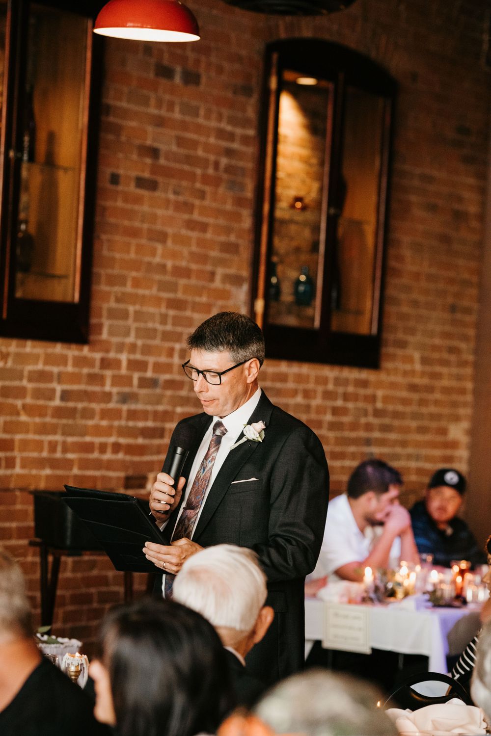 Man in suit giving wedding speech at reception in rustic brick venue.