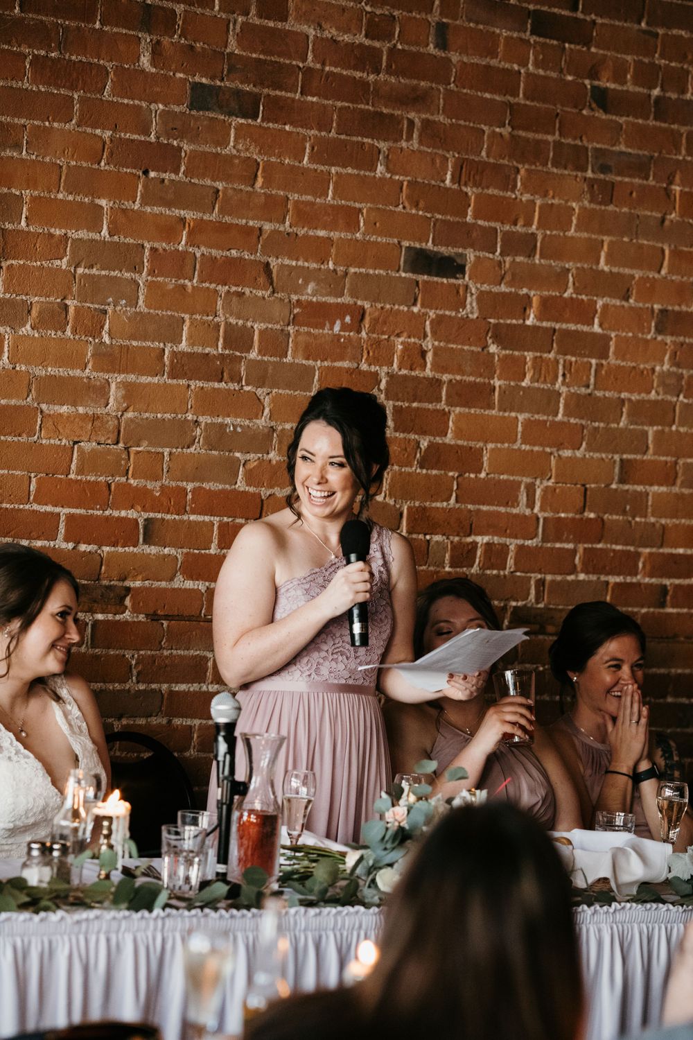 Smiling bridesmaid delivering wedding speech at rustic brick venue reception.