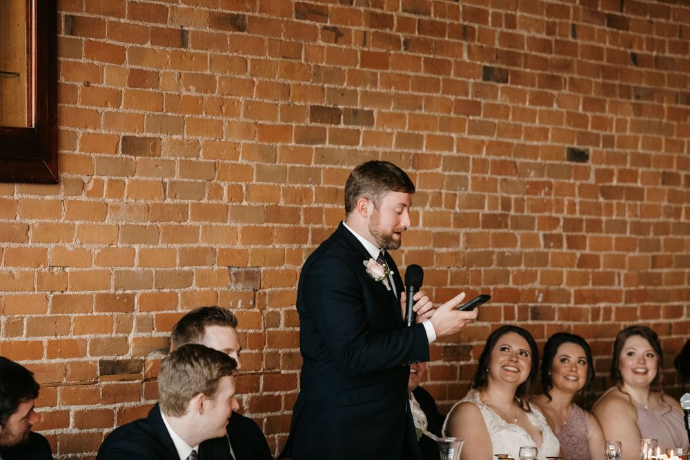 Groom giving speech to smiling guests at rustic wedding reception.