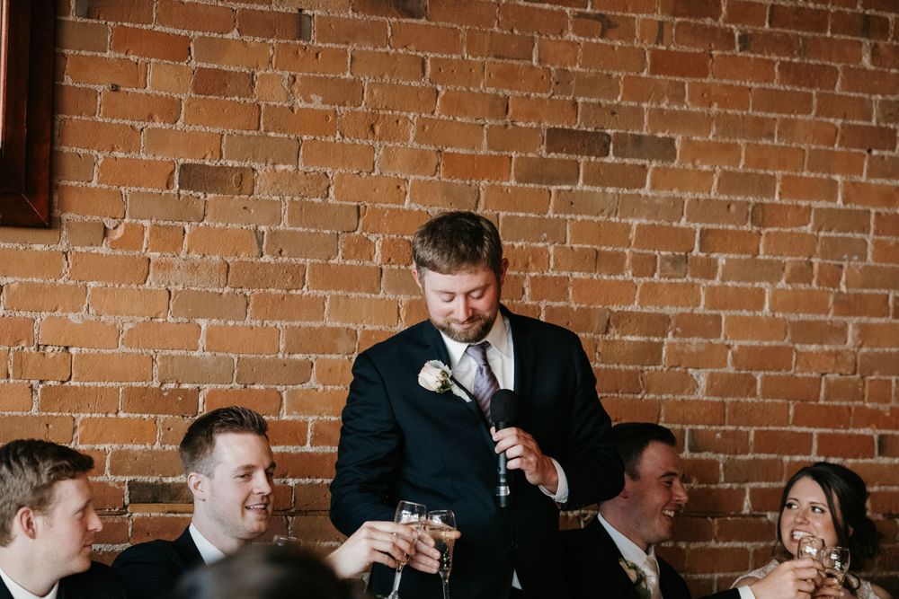 Groom in black suit giving a heartfelt toast at wedding reception in front of exposed brick wall.
