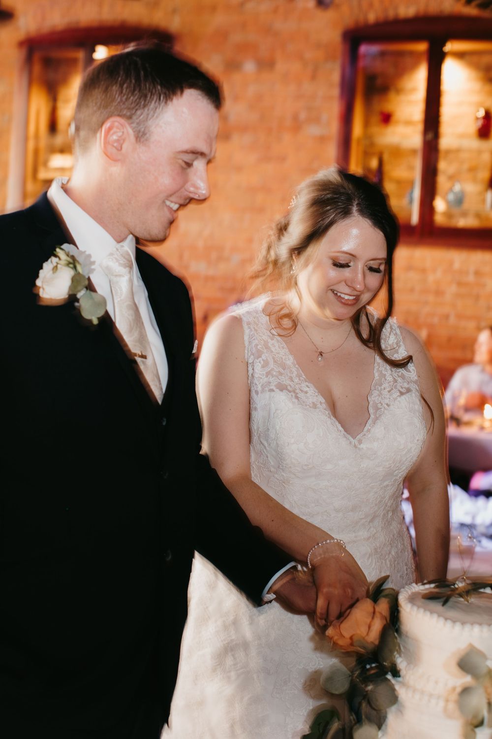 Bride and groom smiling while cutting their white wedding cake at indoor reception venue.