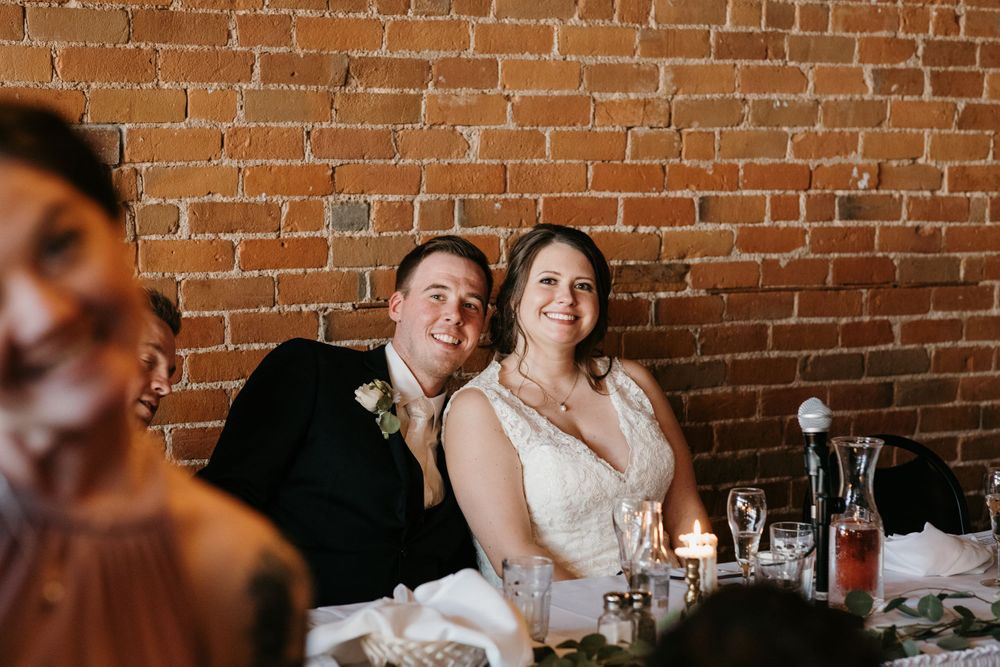 Smiling bride and groom seated at wedding reception table in front of exposed brick wall.