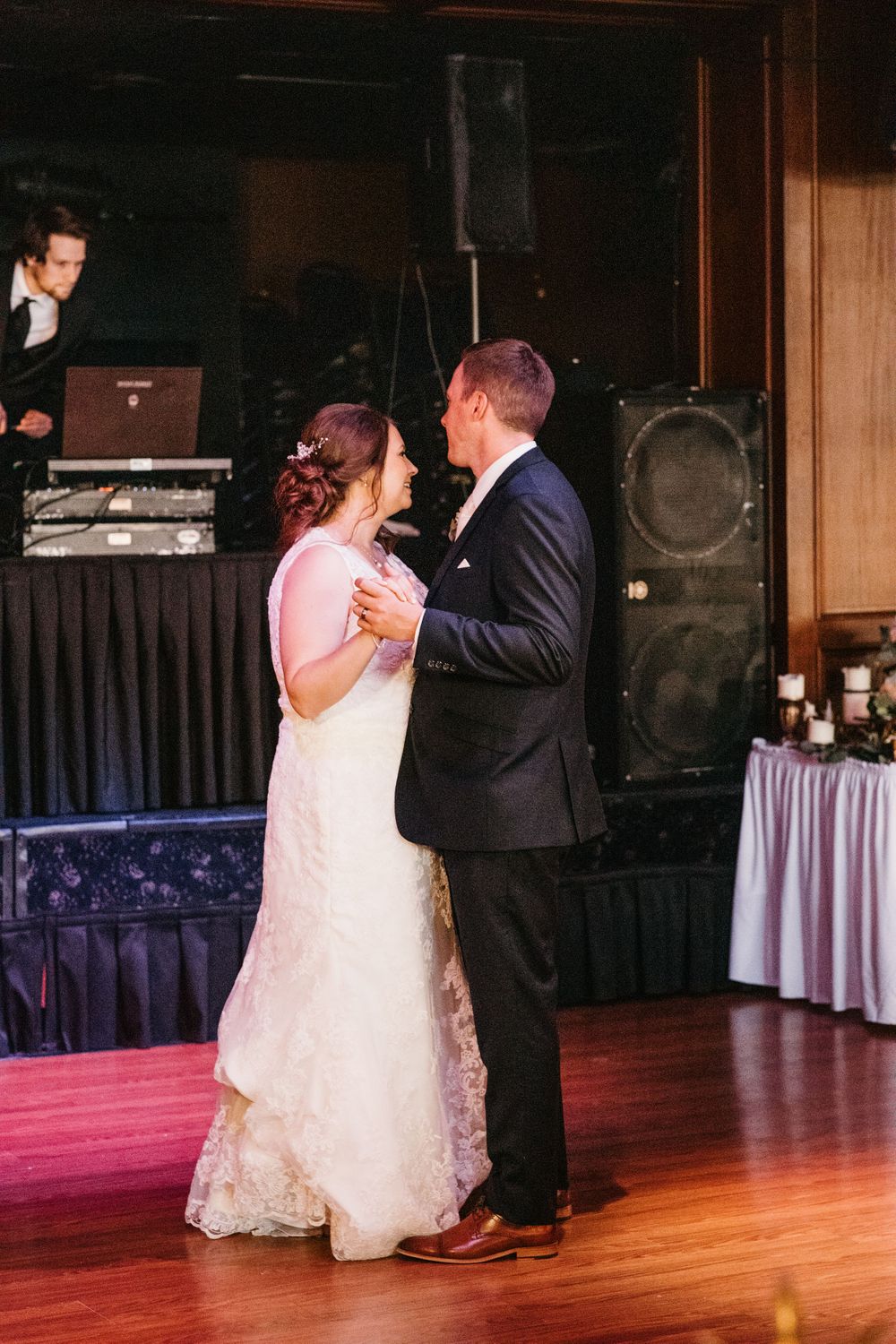 Bride and groom share their first dance at wedding reception with DJ setup visible in background.