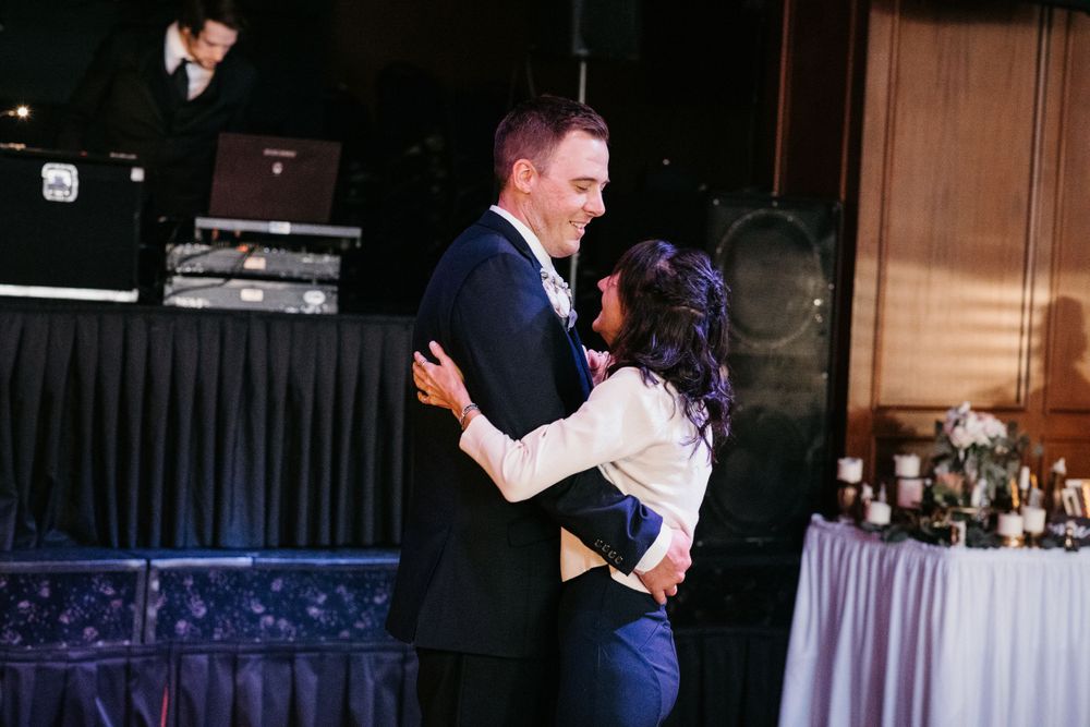 Groom dances with female guest at wedding reception with DJ and floral arrangements visible.