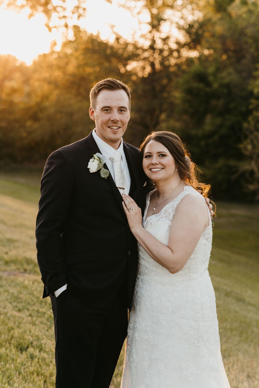 Smiling bride and groom pose together outdoors at golden hour with lush green trees behind them.