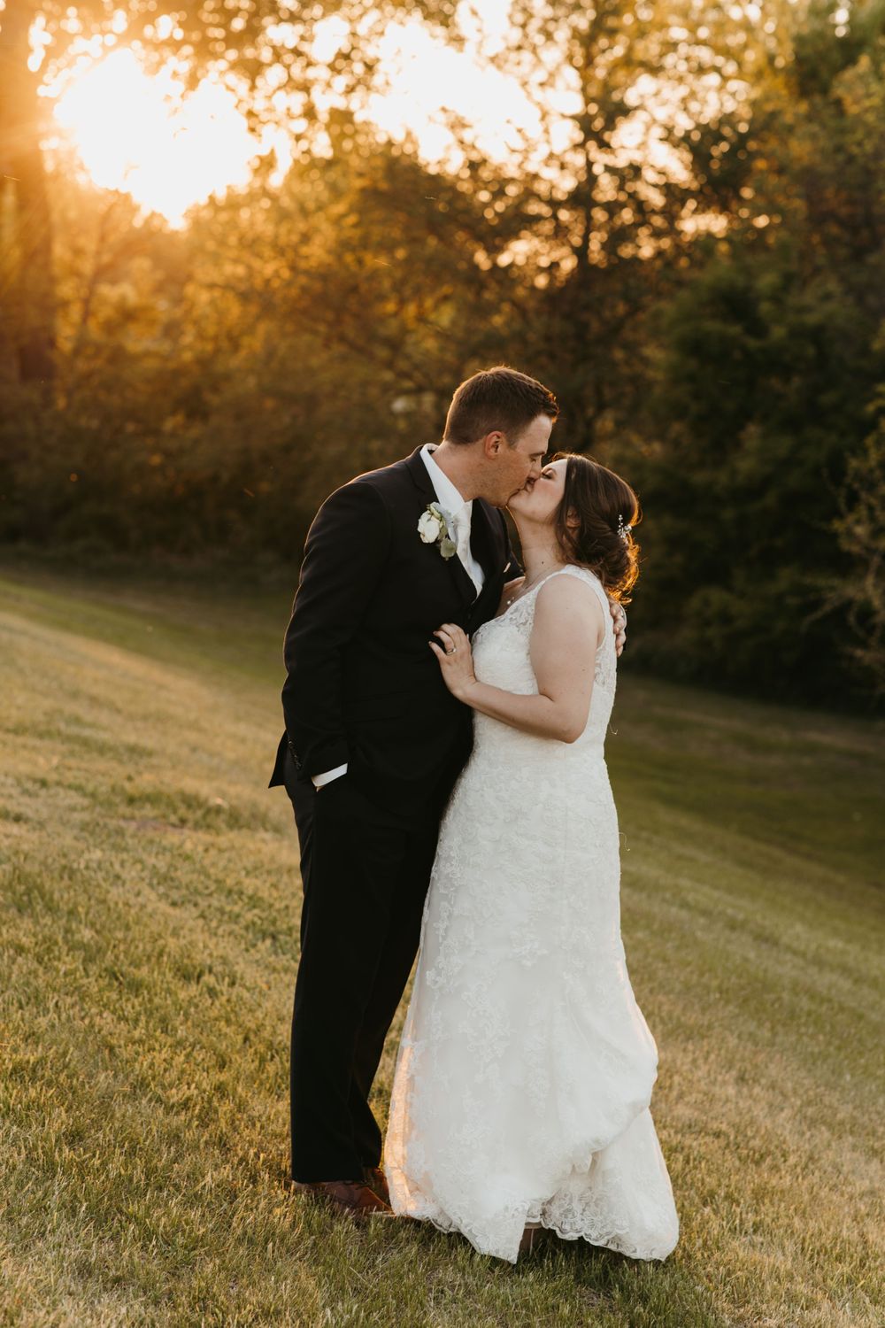 Bride and groom sharing a romantic kiss on green lawn during beautiful golden hour sunset.