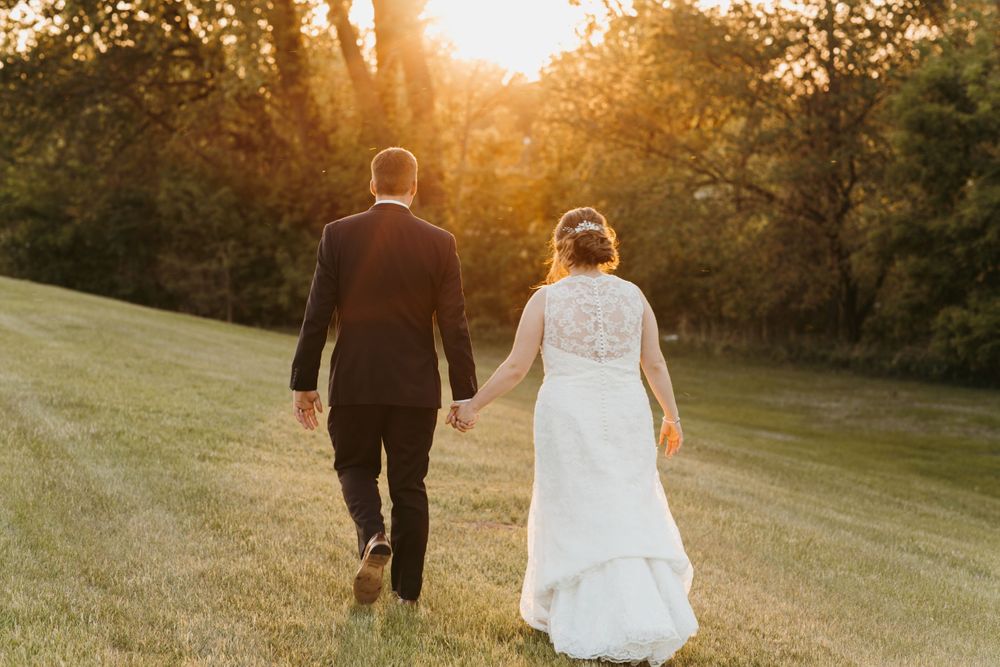 Bride and groom walking hand in hand away from camera across lawn at golden sunset.